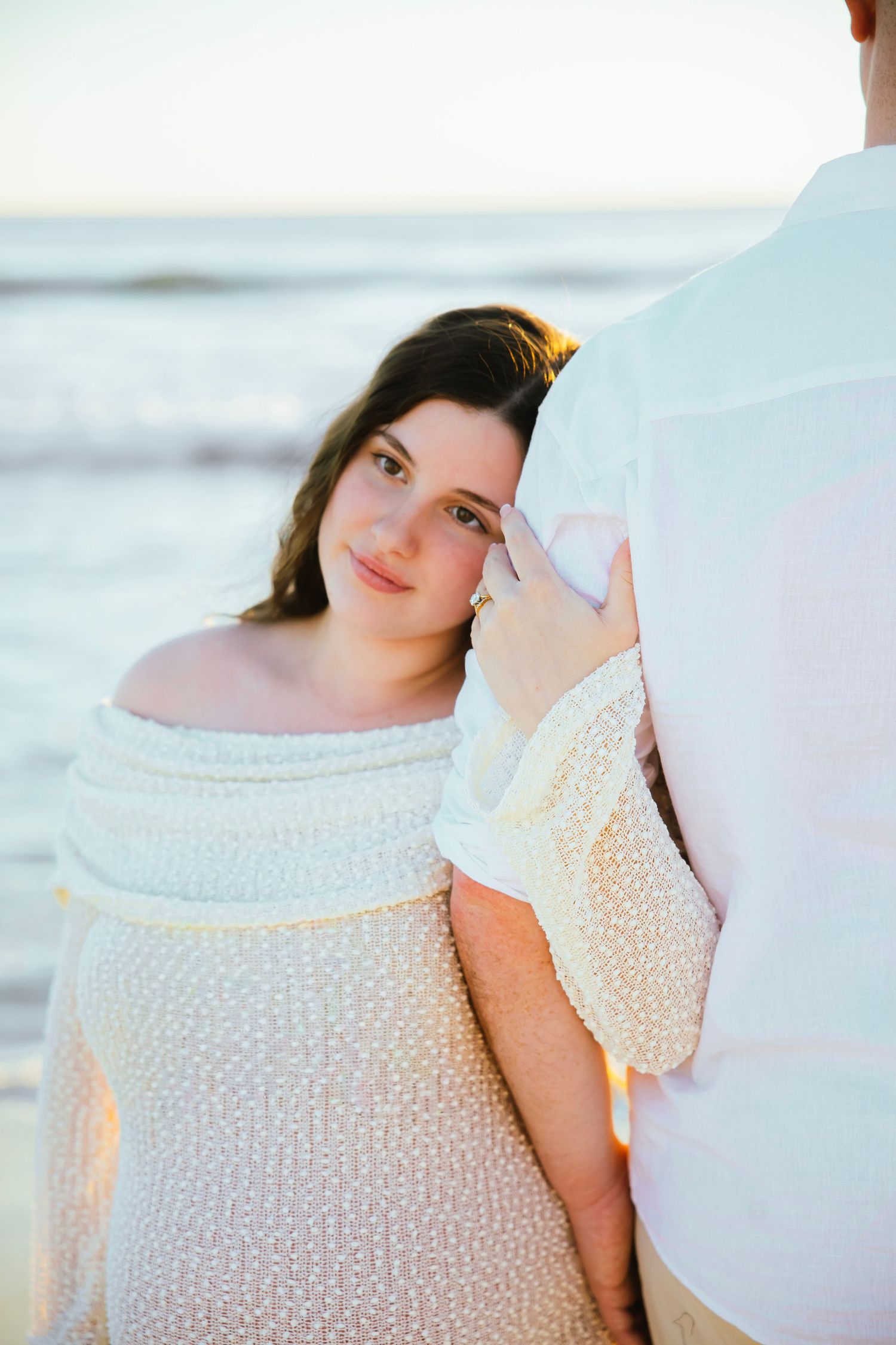 Pregnant woman in off-shoulder white knit dress leaning against partner on beach at golden hour.