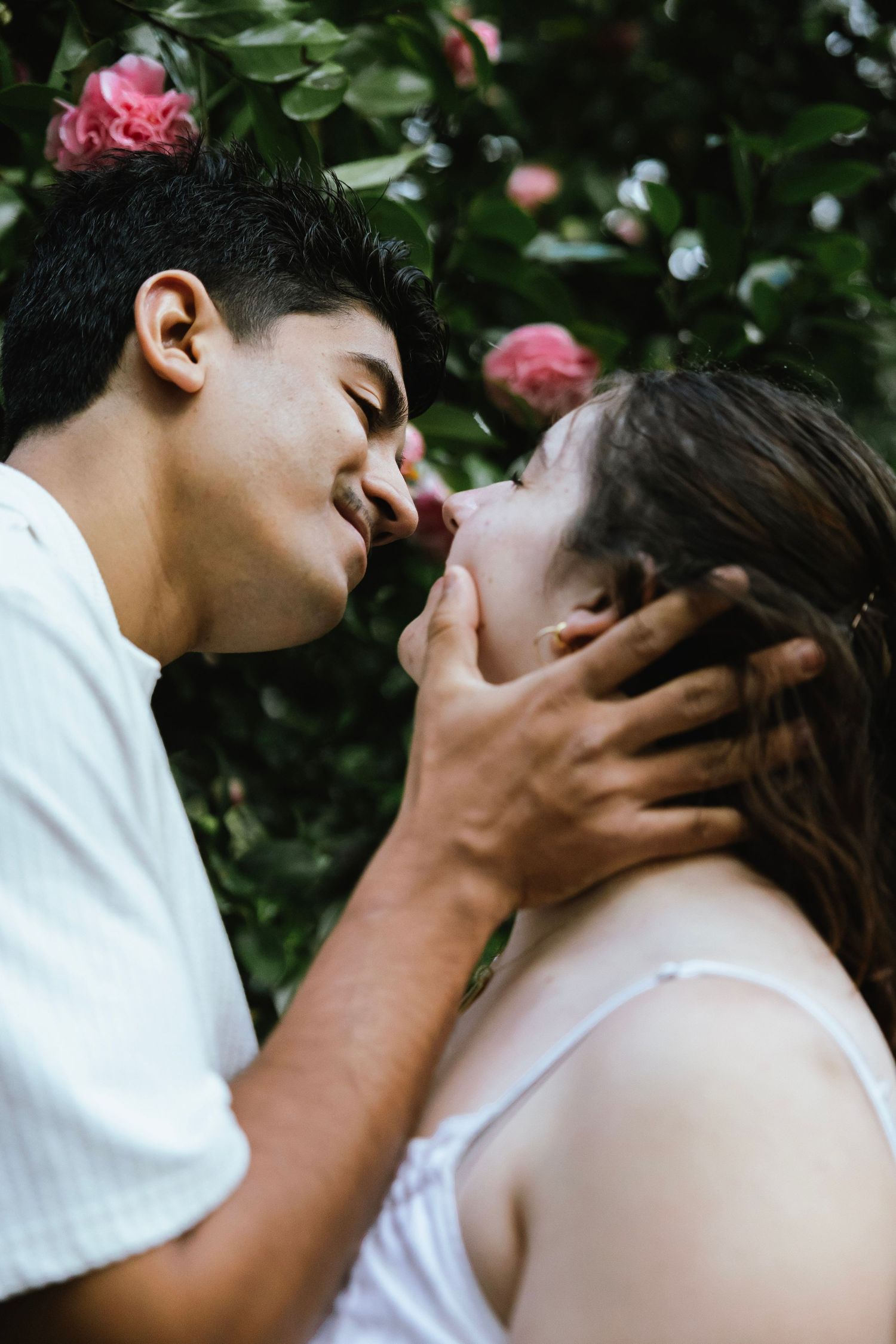 Close-up of a couple sharing a romantic kiss surrounded by green leaves and pink roses.