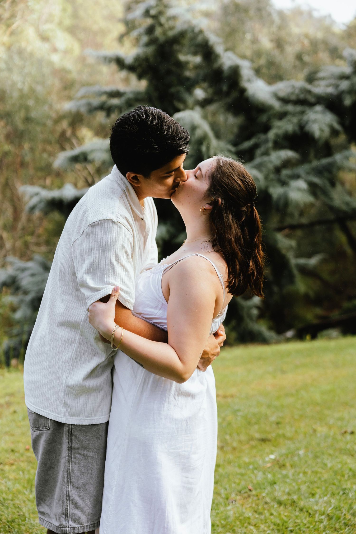 Romantic couple sharing a kiss outdoors with evergreen trees in the background.