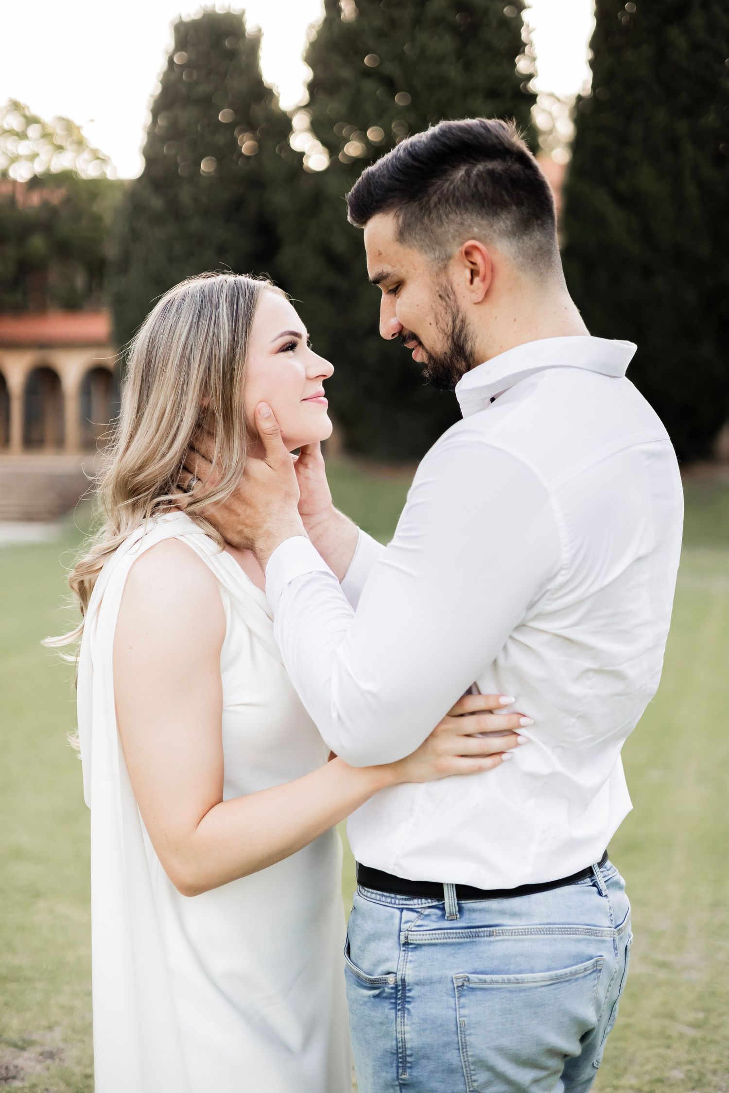 Loving couple in white outfits share an intimate moment outdoors with cypress trees in background.