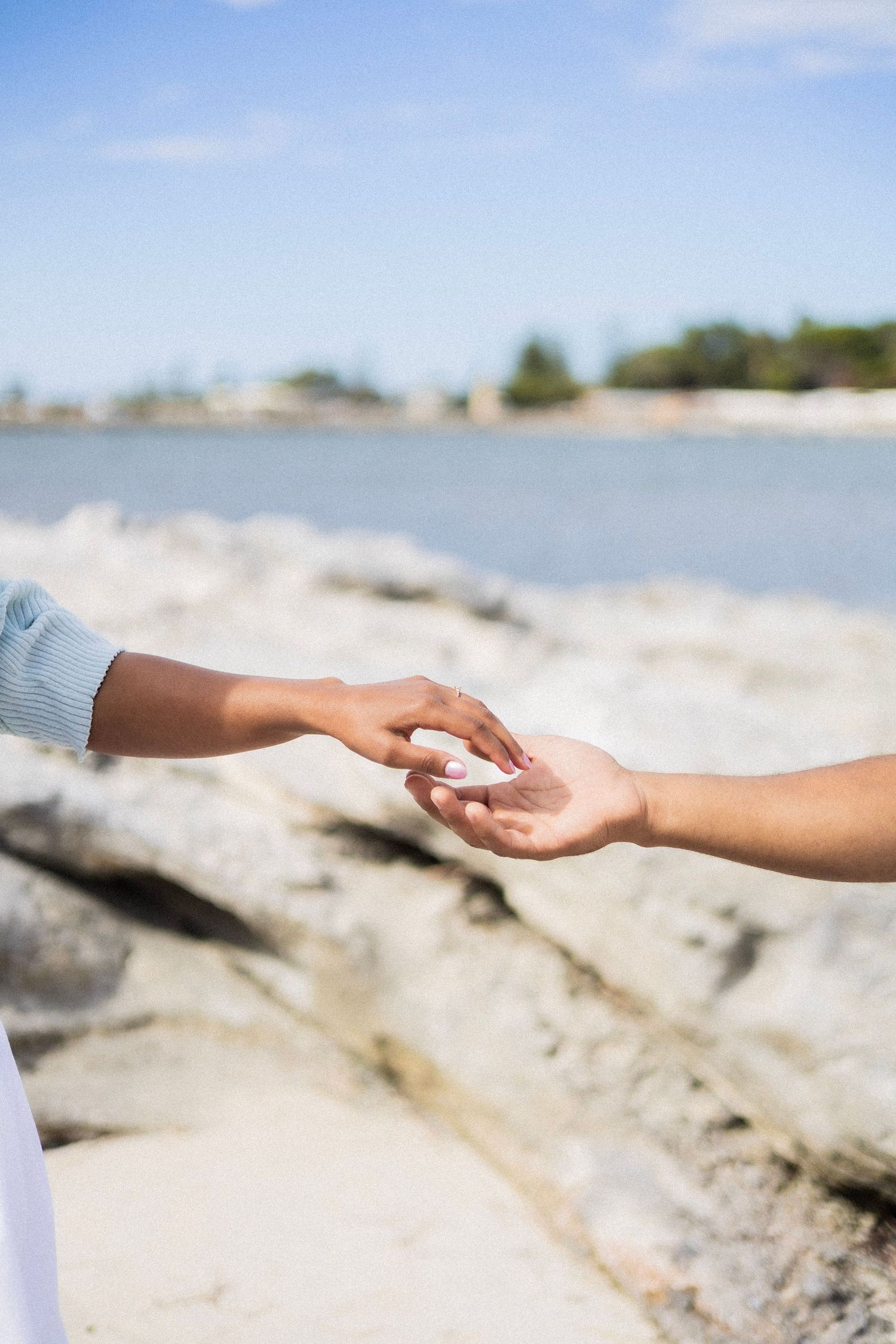 Two hands gently touching fingertips on a rocky beach with ocean and blue sky in the background.