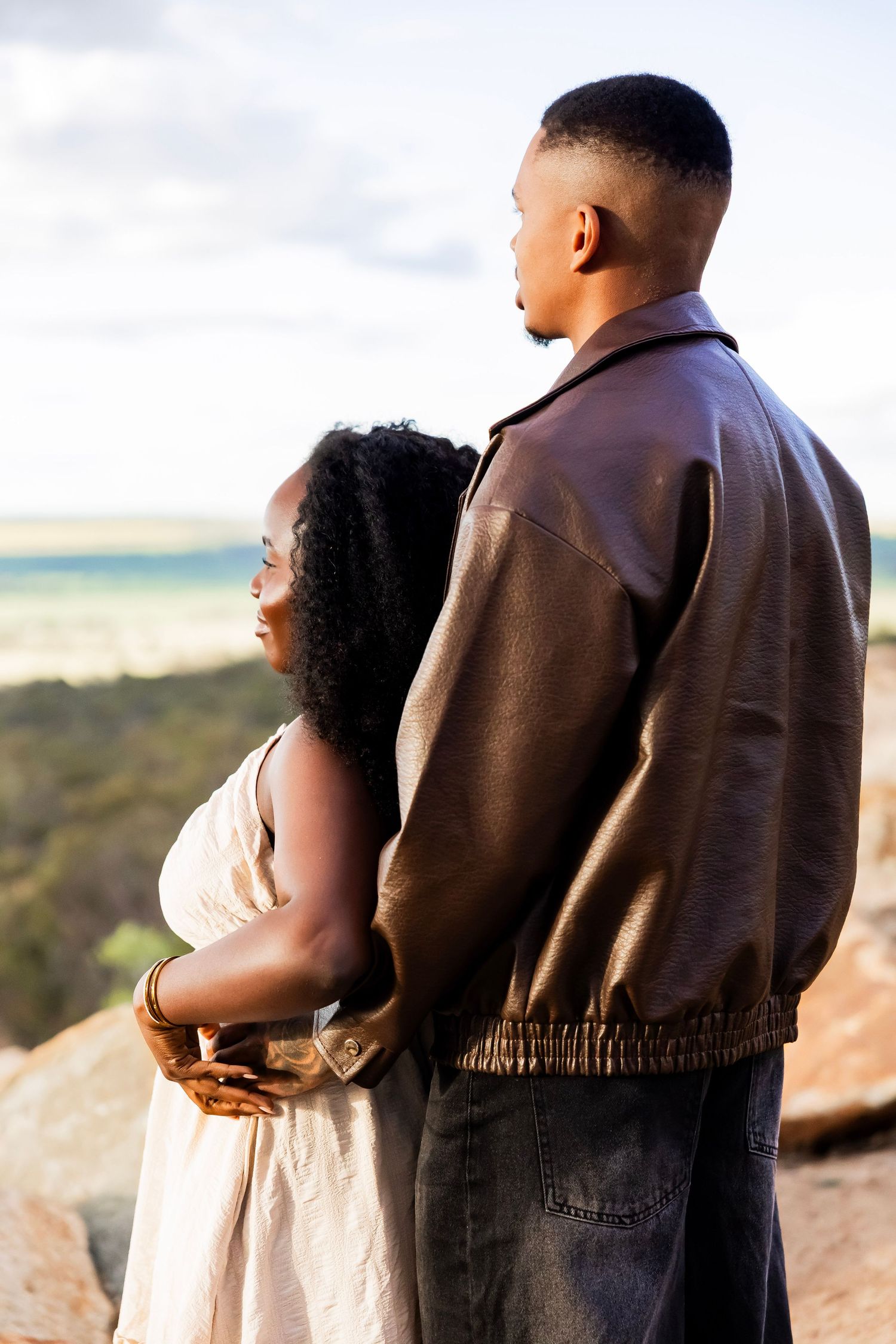 A Black couple stands together on rocky terrain, the man in a leather jacket embracing the woman in a white dress, gazing at a scenic view.