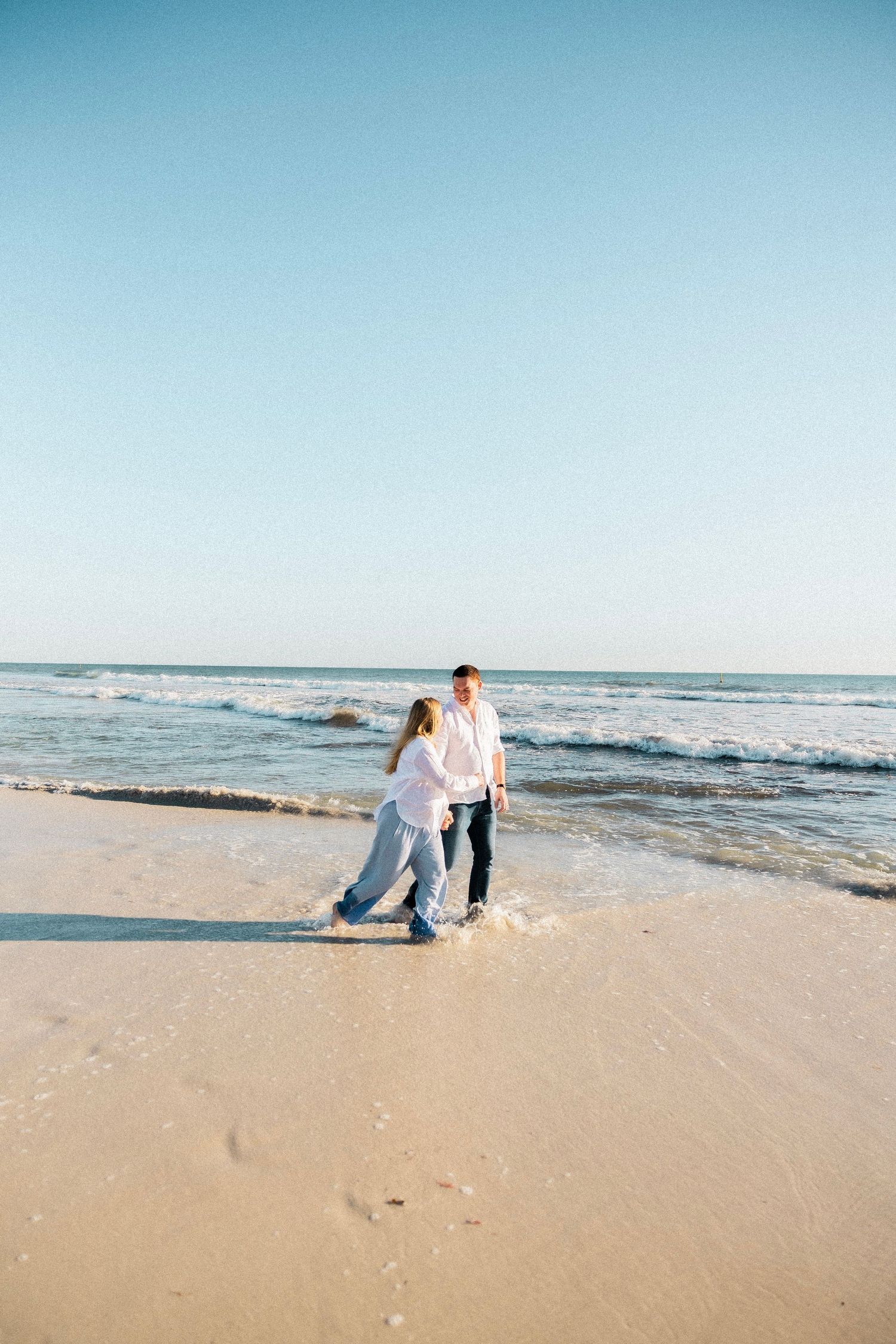 Pregnant couple walking hand in hand along a sunny sandy beach with gentle ocean waves.