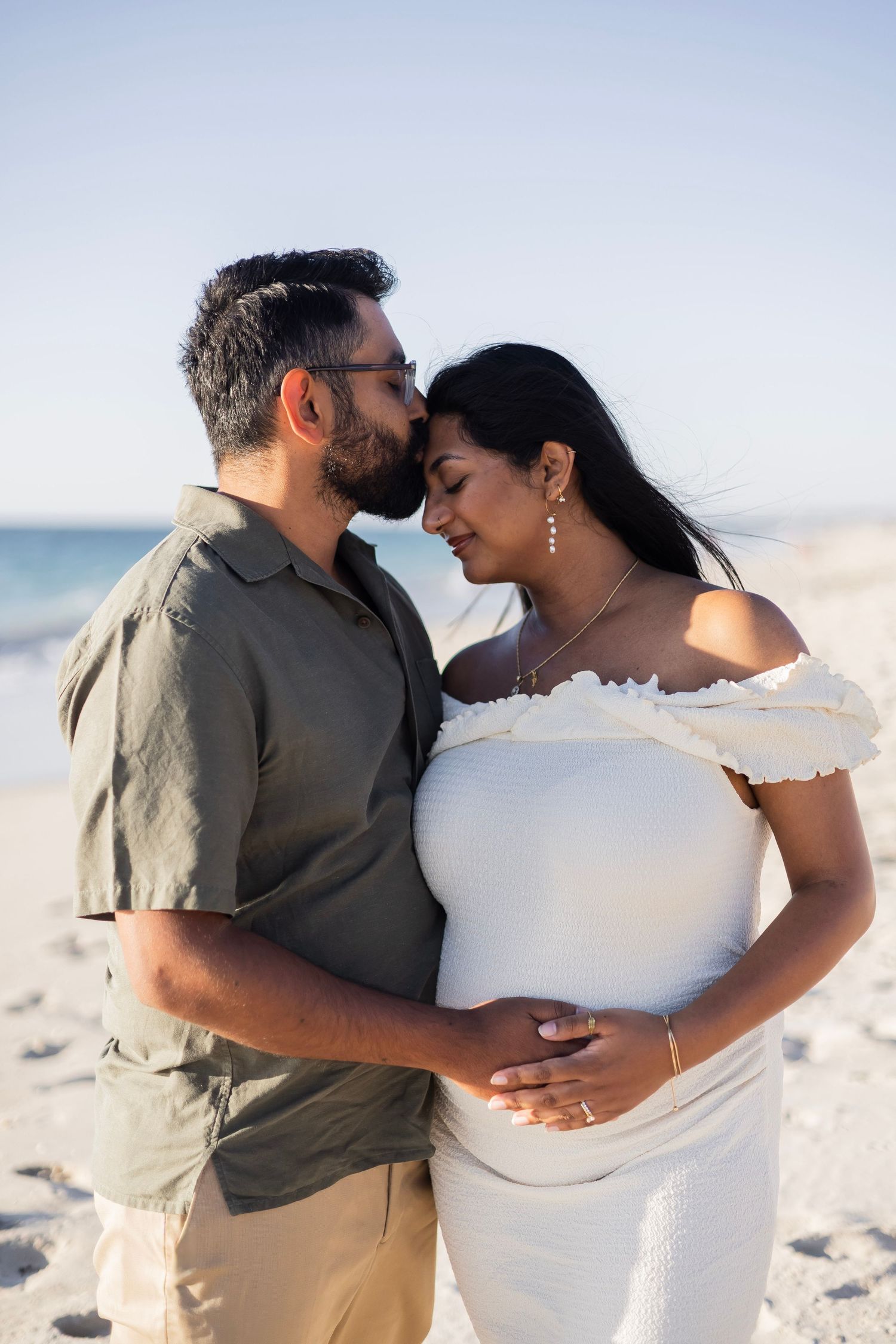 Couple sharing a tender moment on a sunny beach, man kissing pregnant woman's forehead while cradling her baby bump.