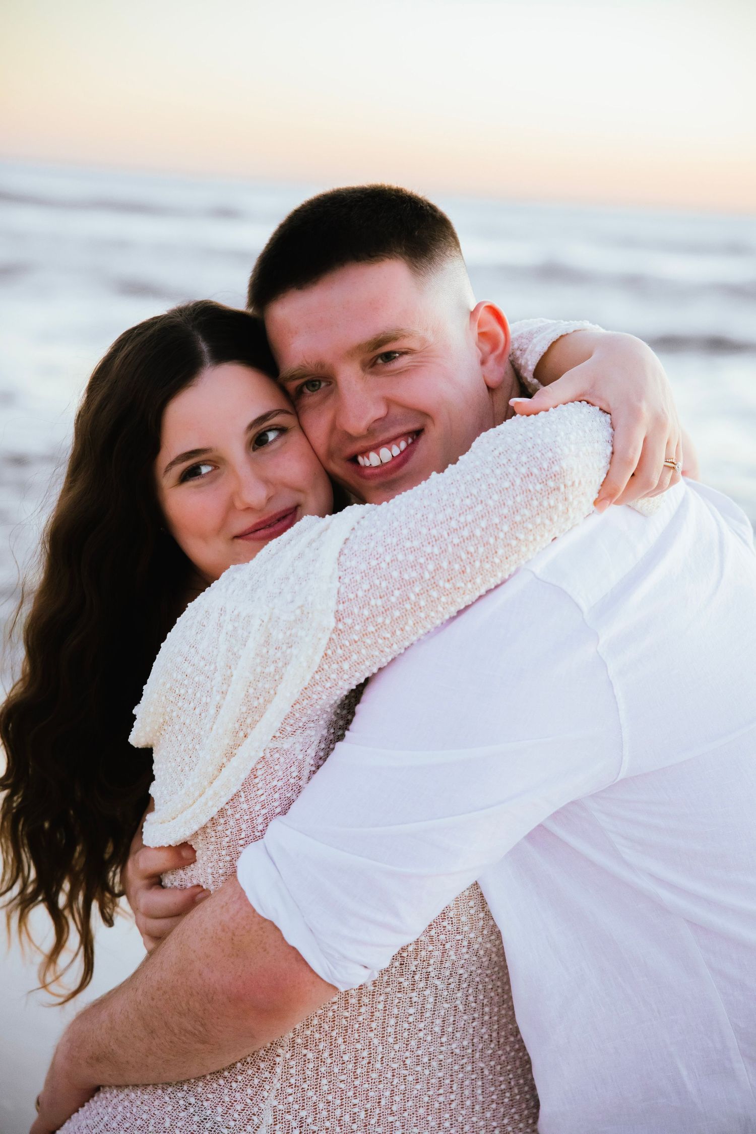 Happy couple hugging on beach at sunset, smiling man in white shirt embraced by woman in white lace dress.