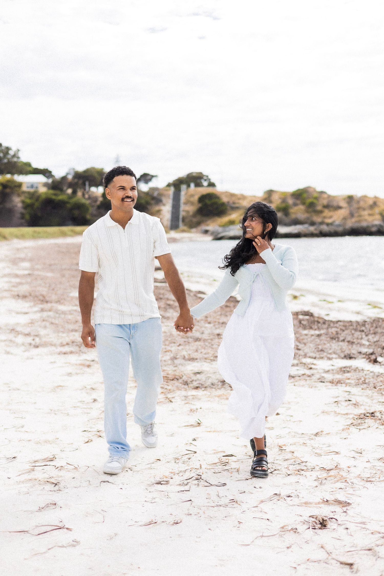 A happy couple walks hand in hand along a sandy beach with coastal scenery and water behind them.