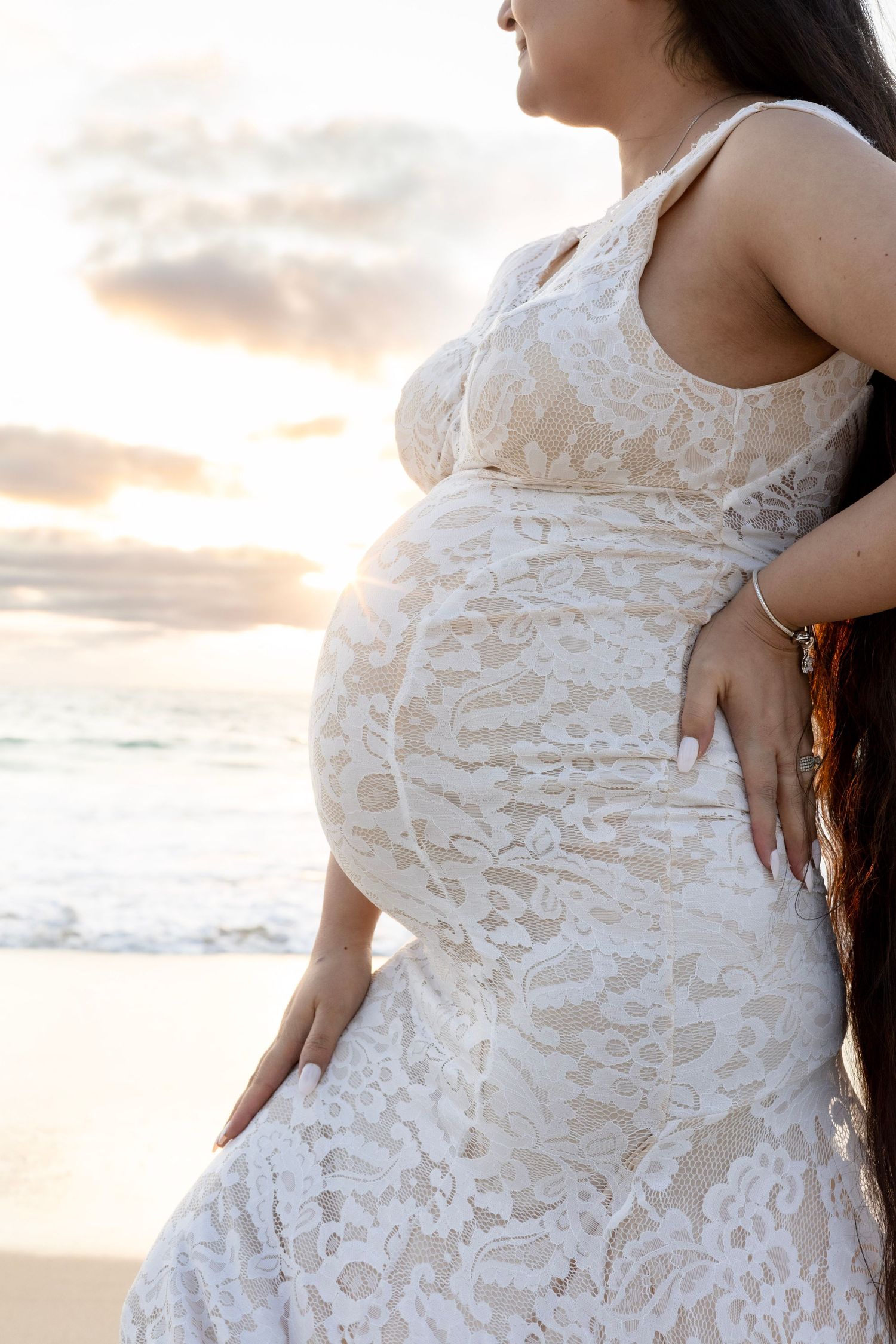 Close-up of pregnant woman in white lace dress cradling baby bump at beach sunset.