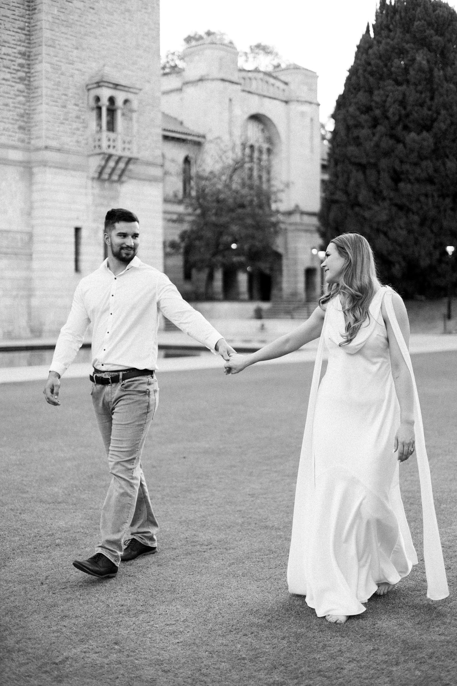 Black and white photo of couple holding hands walking near historic stone building with cypress tree.