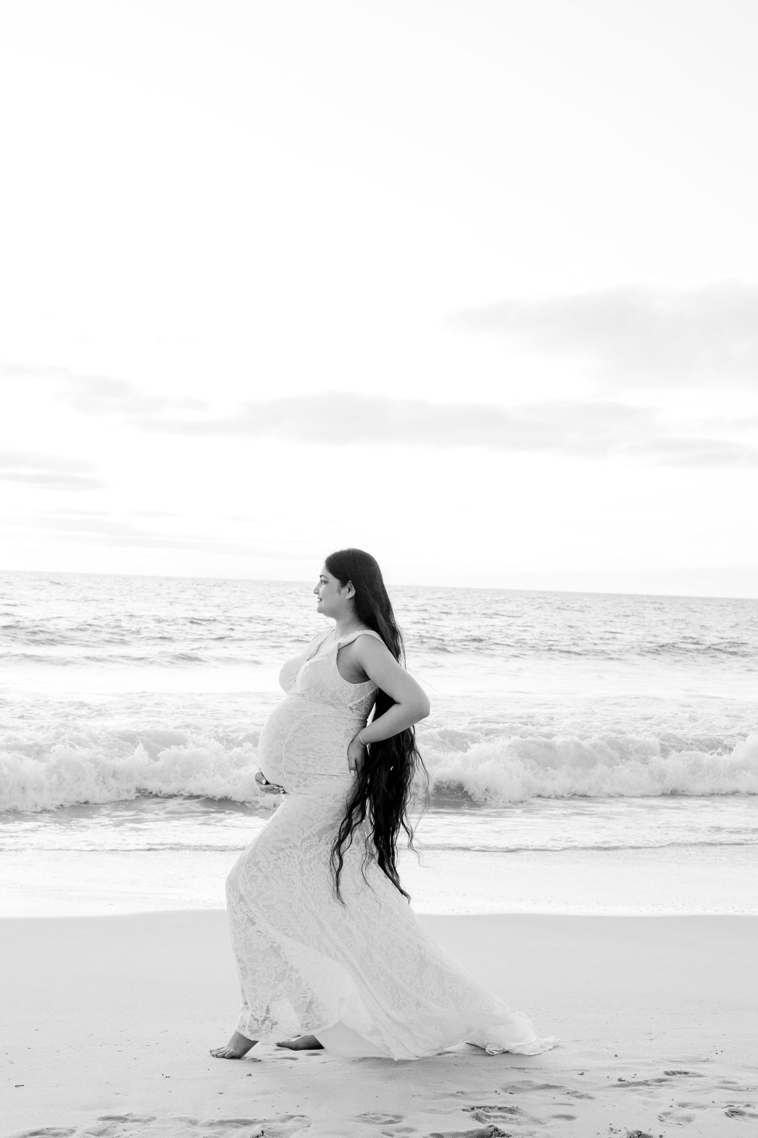 Black and white side profile of pregnant woman in flowing white lace dress standing on beach at sunset.