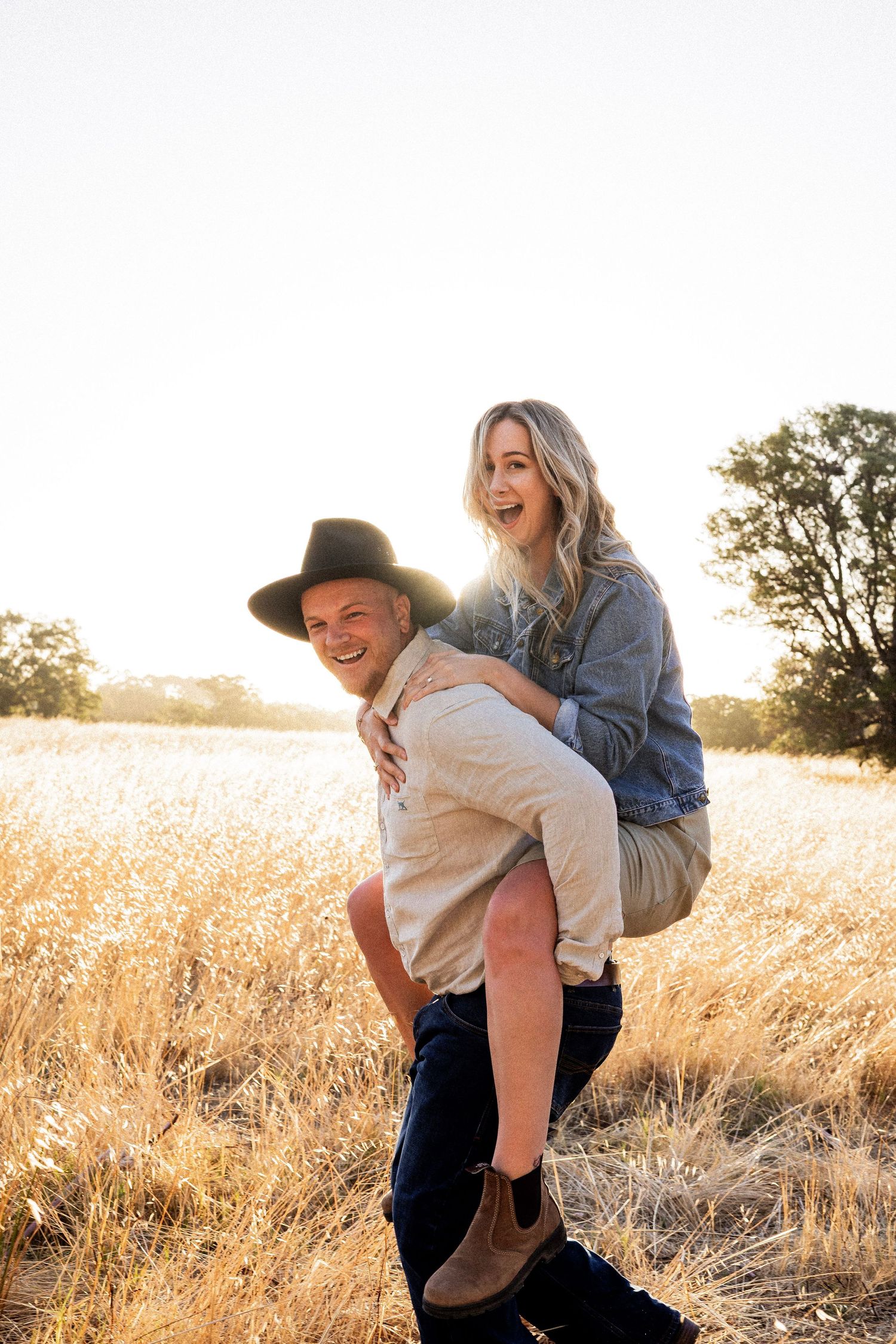 A smiling woman gets a piggyback ride from a man in a wide-brim hat in a golden sunlit field.