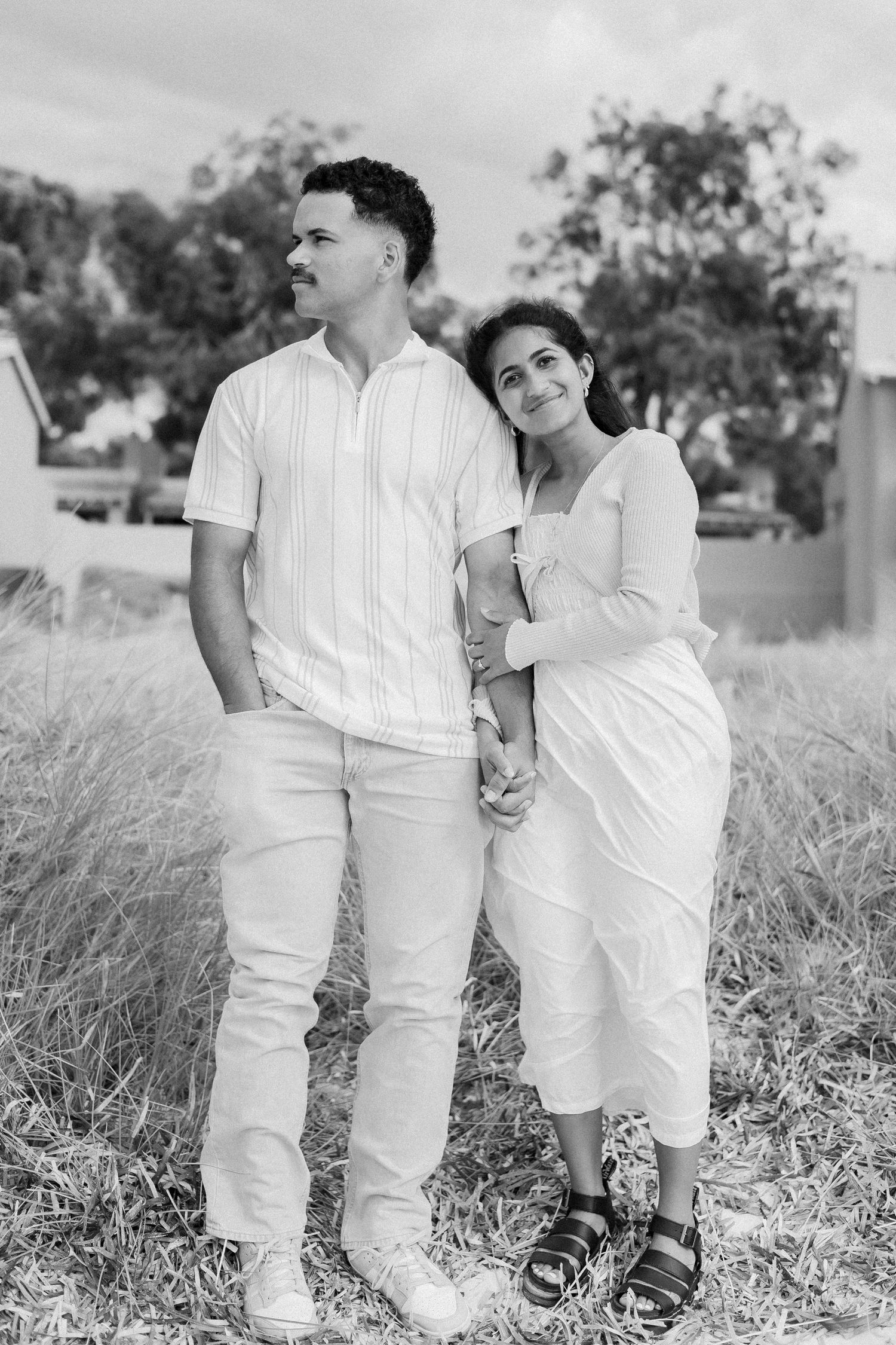 Black and white portrait of couple standing in field, woman leaning on mans shoulder holding hands.