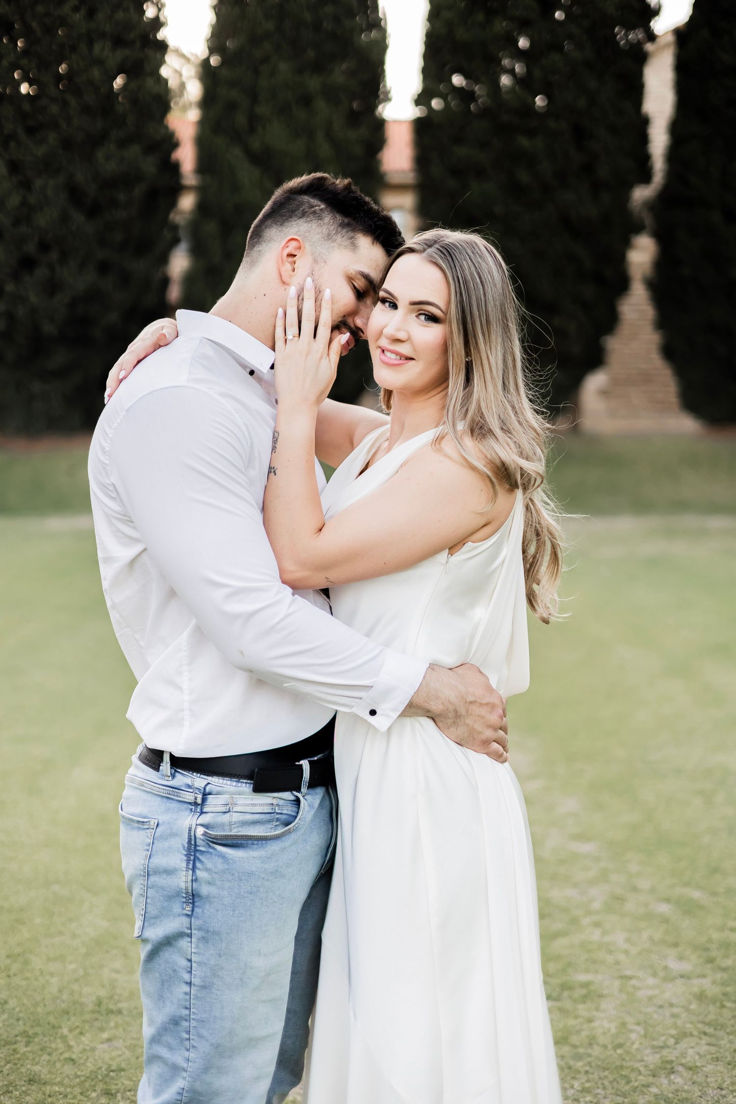 Couple embraces tenderly in a garden, woman in white dress smiling as man holds her face near cypress trees.