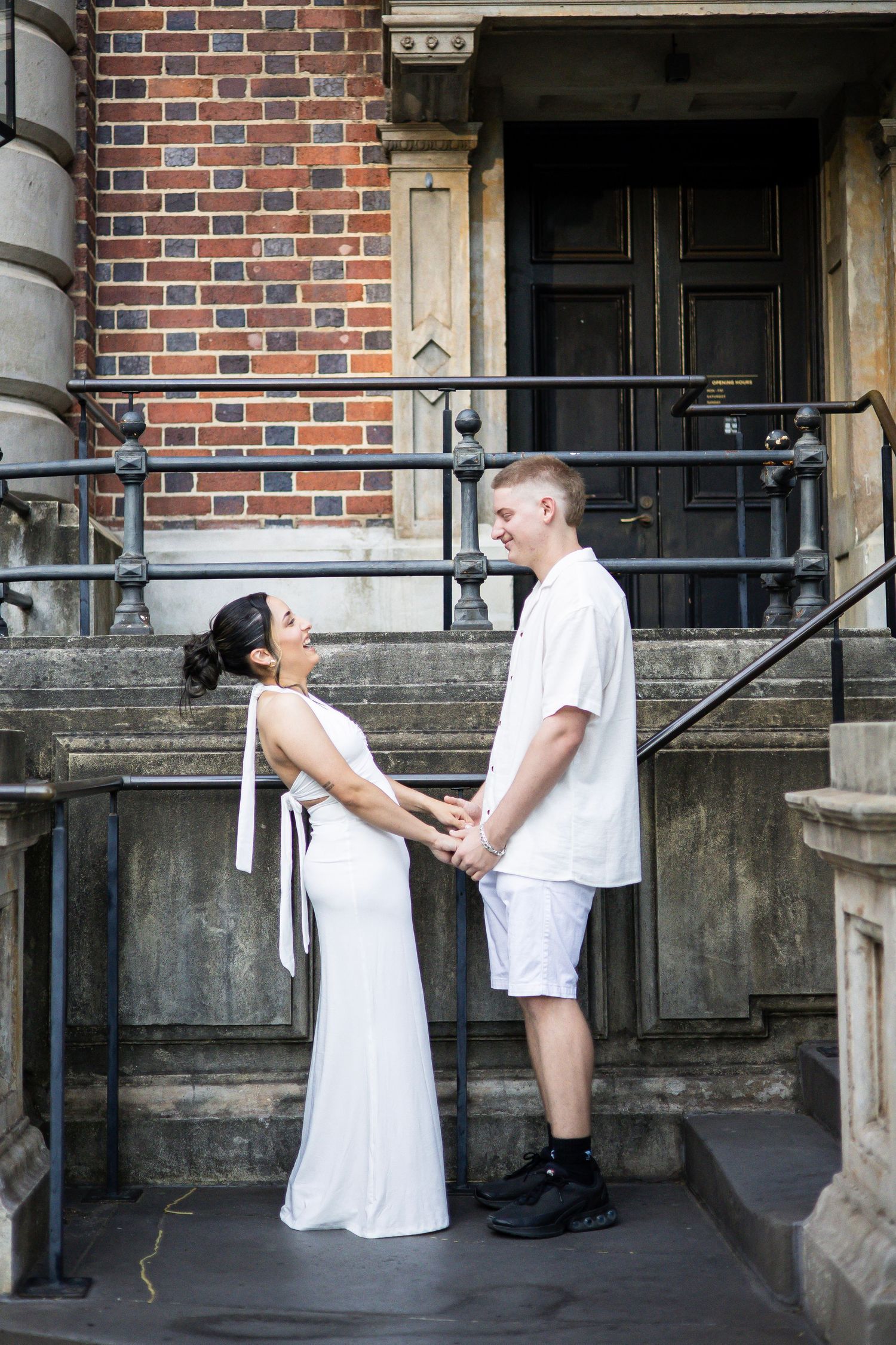 Smiling couple in white outfits holding hands on historic stone steps with brick building backdrop.