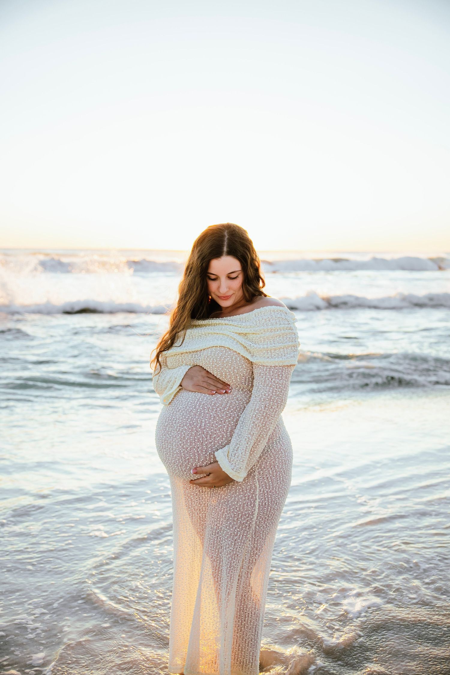 Pregnant woman in white knit dress cradling her baby bump on a beach at sunset with ocean waves behind her.