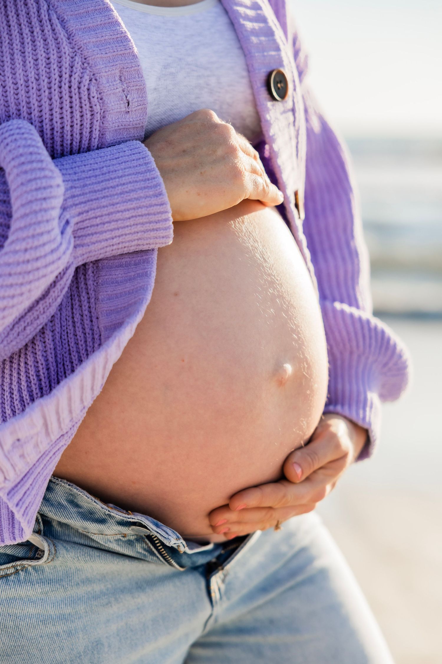 Close-up of a pregnant woman's bare belly held by her hands, wearing a purple cardigan and jeans.
