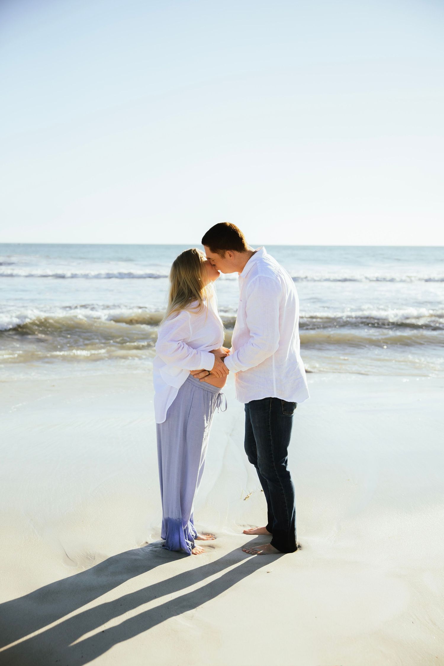 A couple shares a tender kiss on a sunny beach, the woman showing a pregnant belly in a flowing lavender skirt.