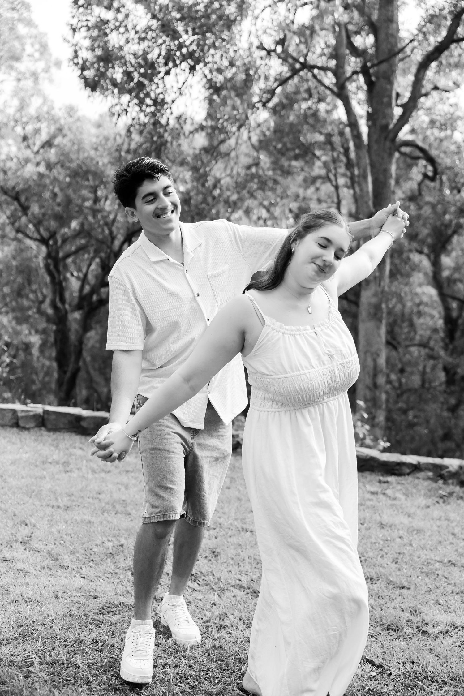 A happy couple dances outdoors in black and white, the man spinning the woman in a white dress among trees.