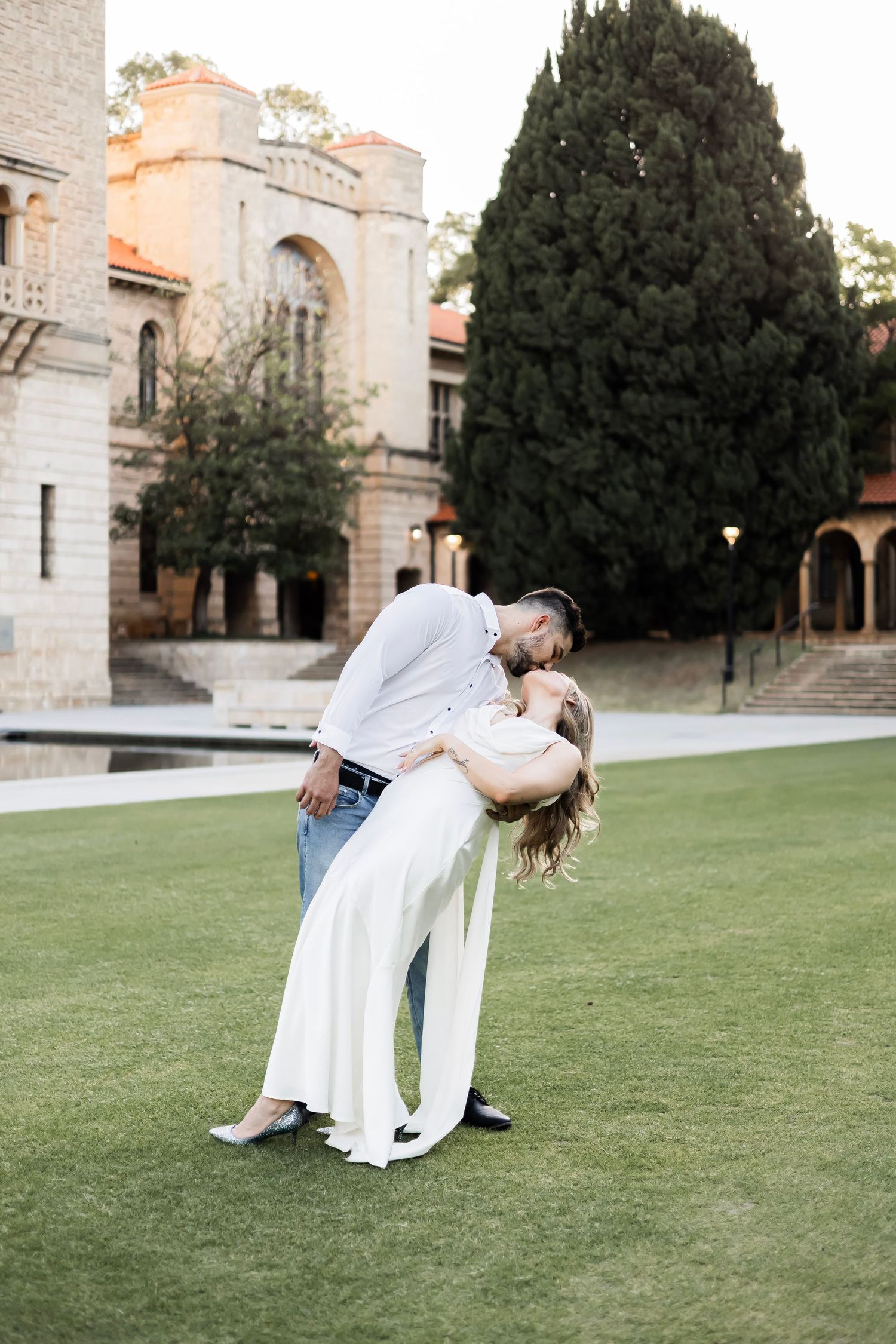 Groom dipping bride in elegant white dress for a romantic kiss on green lawn near historic stone building.