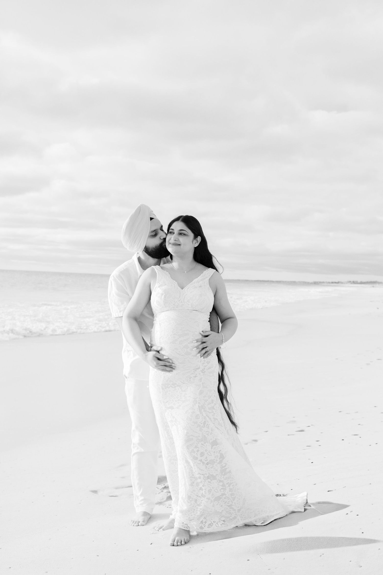Black and white photo of pregnant couple embracing on beach, man in turban kissing woman's head.