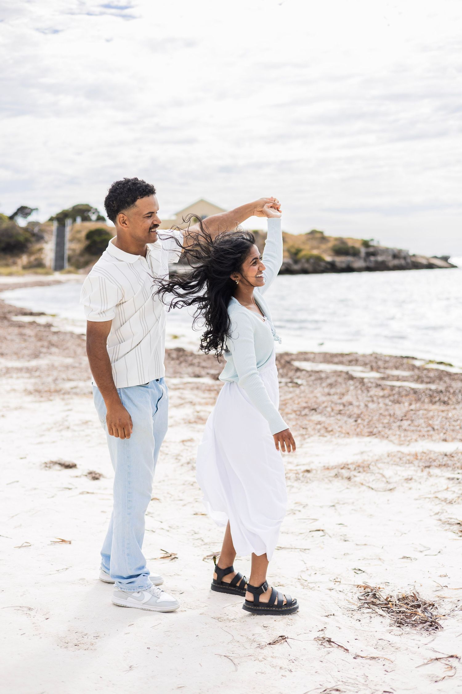 A joyful couple dances on a white sand beach near the ocean on a cloudy day.