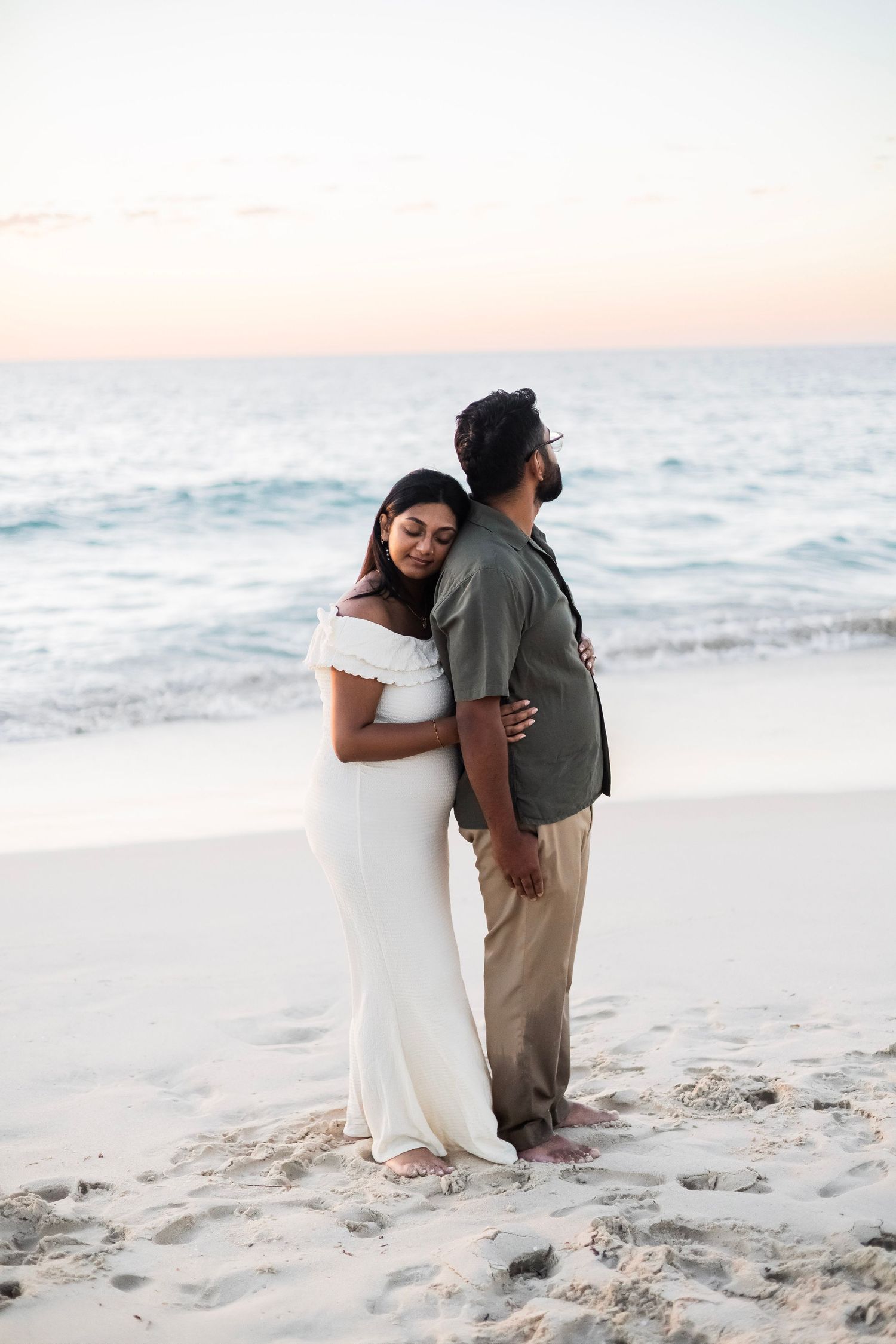 Romantic couple embracing on sandy beach at sunset, woman in white dress leaning against man in green shirt.
