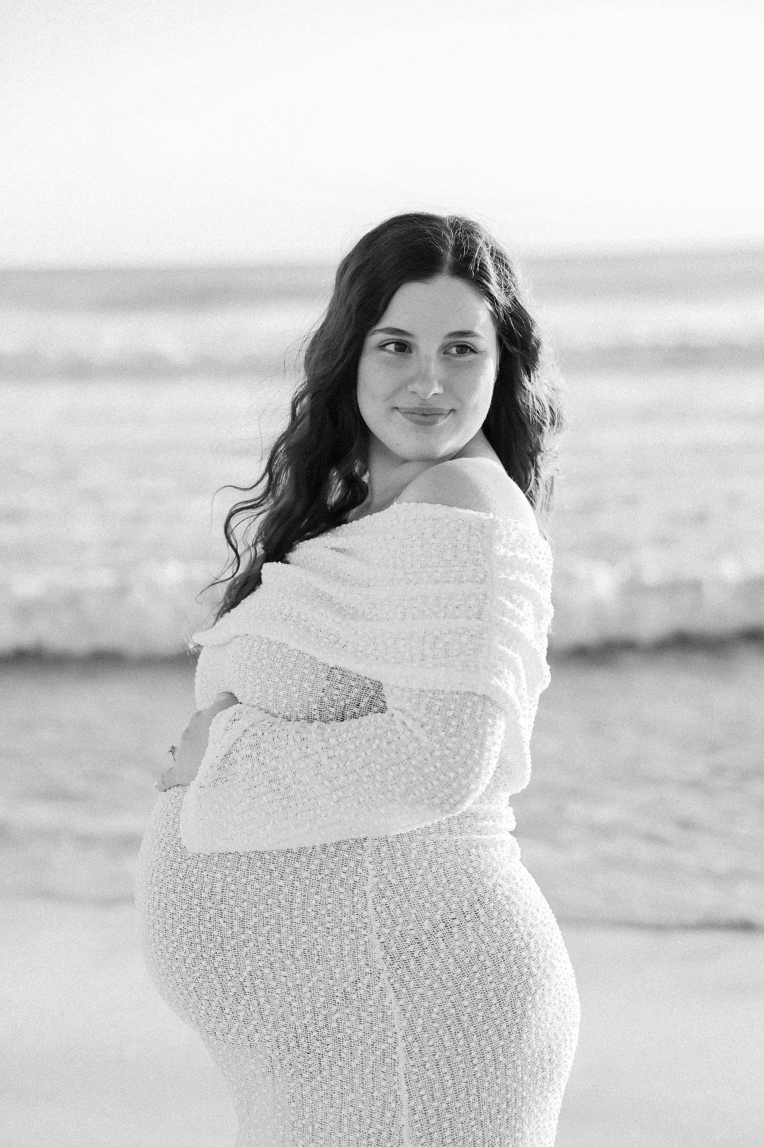 Black and white maternity photo of a pregnant woman in a lace dress posing on a beach with ocean waves behind her.