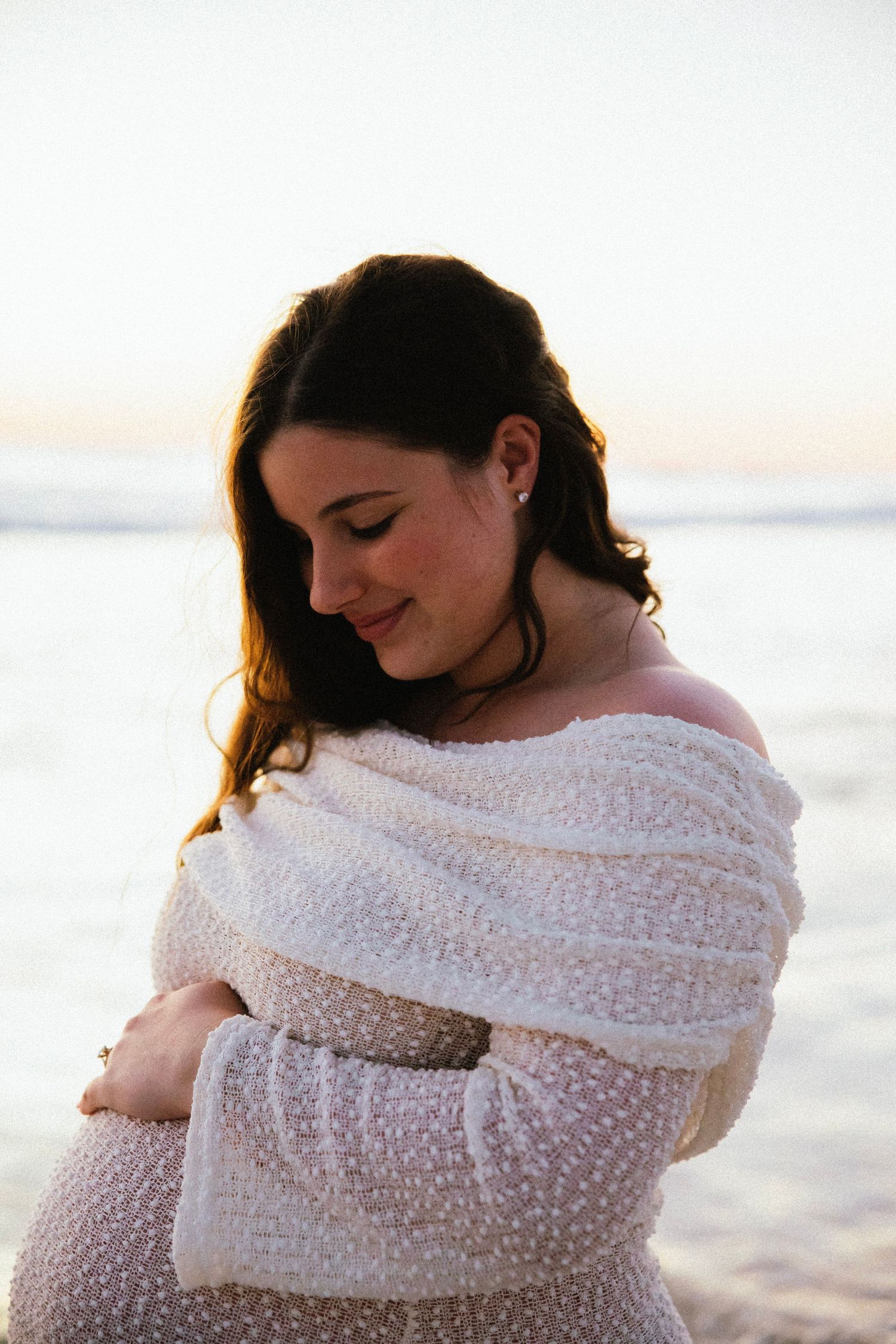 Glowing pregnant woman in off-shoulder white lace dress smiling softly at sunset beach.
