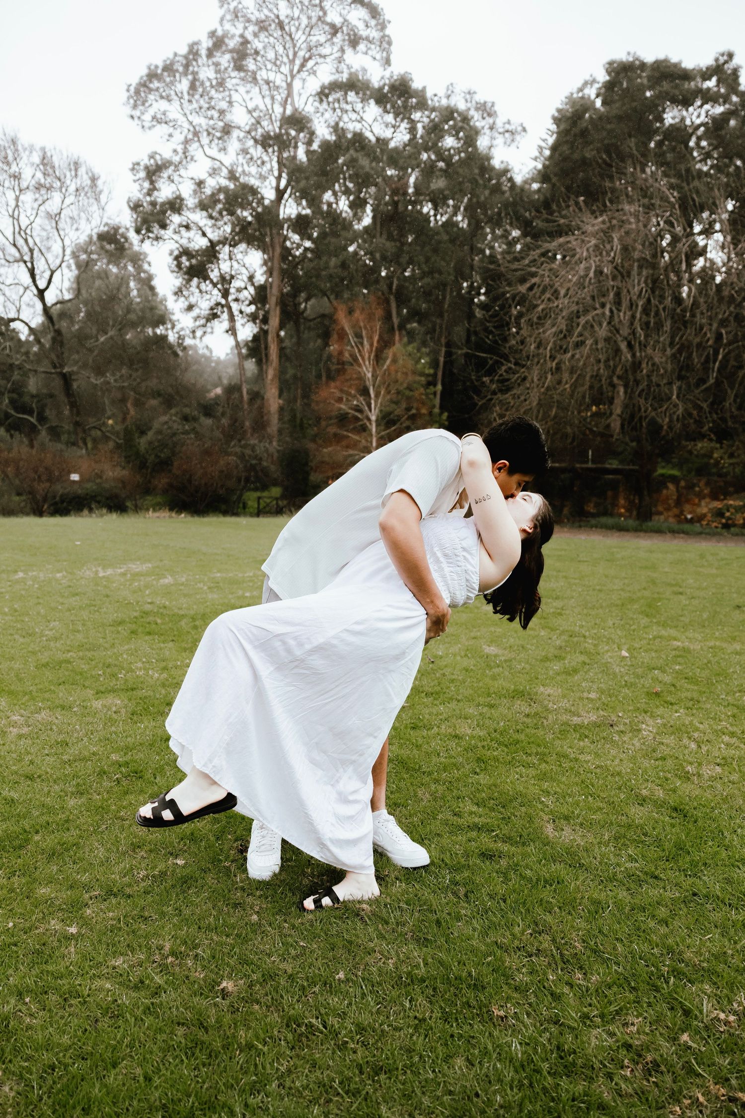 Couple sharing a romantic dip kiss on green lawn with trees in background, bride in white dress.