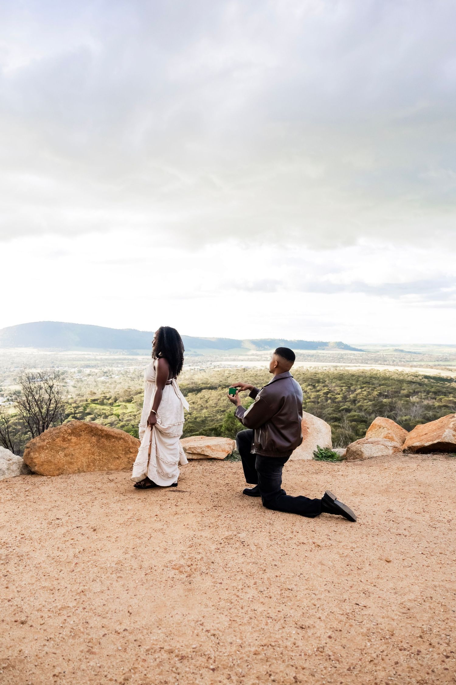 Man proposing on one knee to woman on scenic overlook with mountain valley views under cloudy sky.