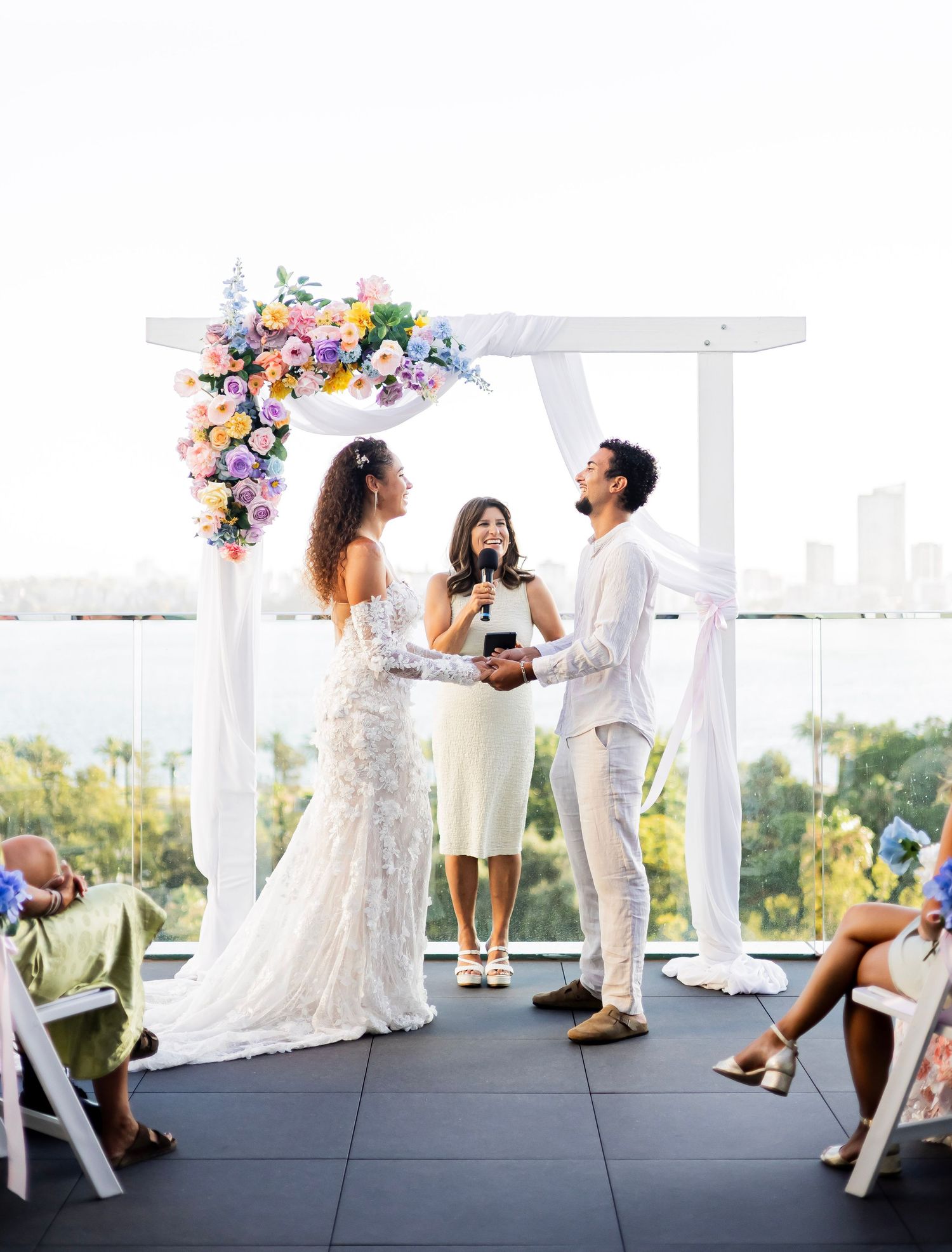 Couple exchanges vows under floral arch with colorful blooms on rooftop with city skyline.