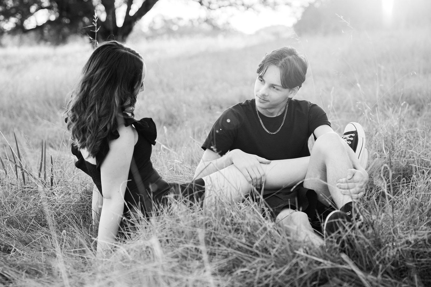 Black and white photo of a young couple sitting together in a sunlit grassy field, gazing at each other.