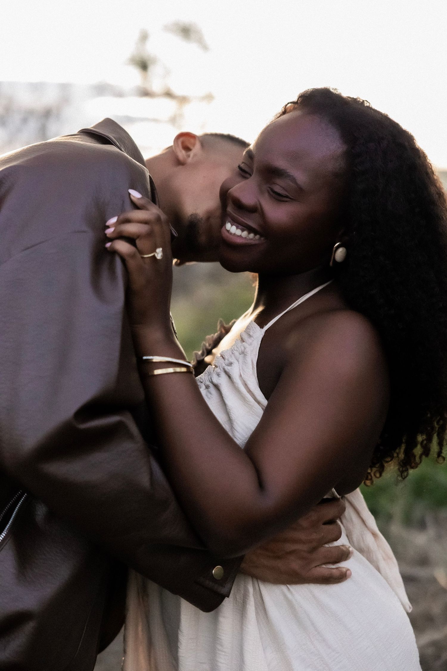 A joyful Black woman in a white dress laughs as her partner kisses her shoulder during a romantic outdoor couple session.