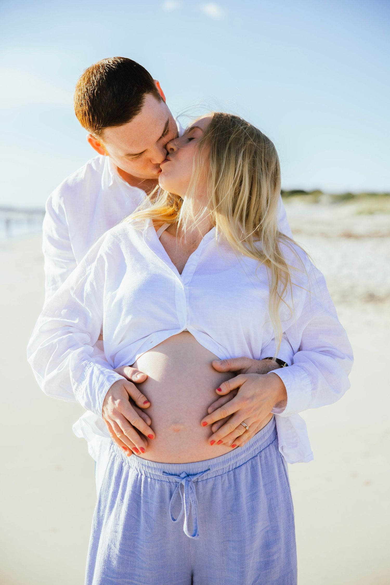 Couple kissing on sunny beach, man tenderly holding pregnant woman's bare belly outdoors.