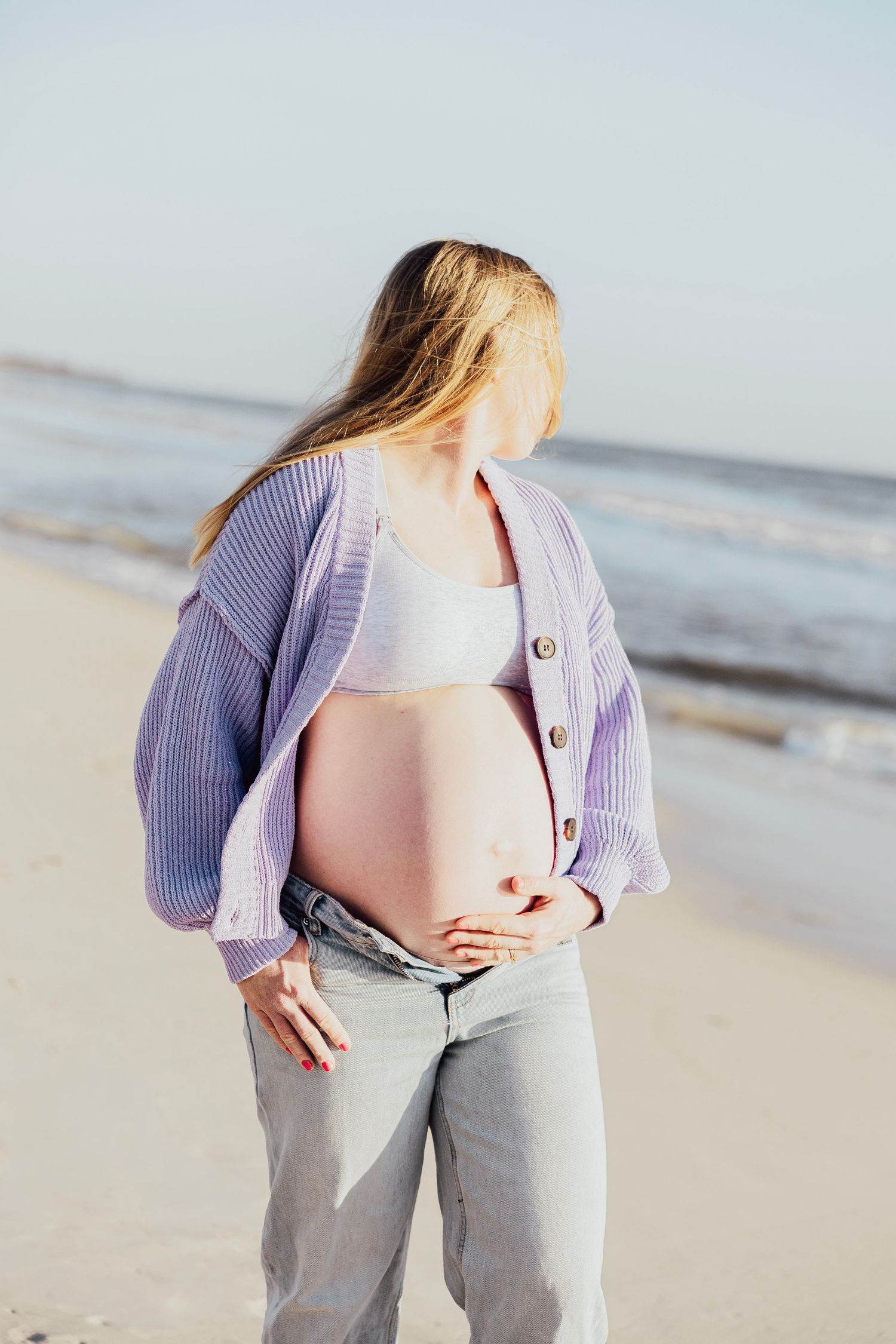 Pregnant woman in purple cardigan and jeans cradles baby bump on a bright sunny beach.