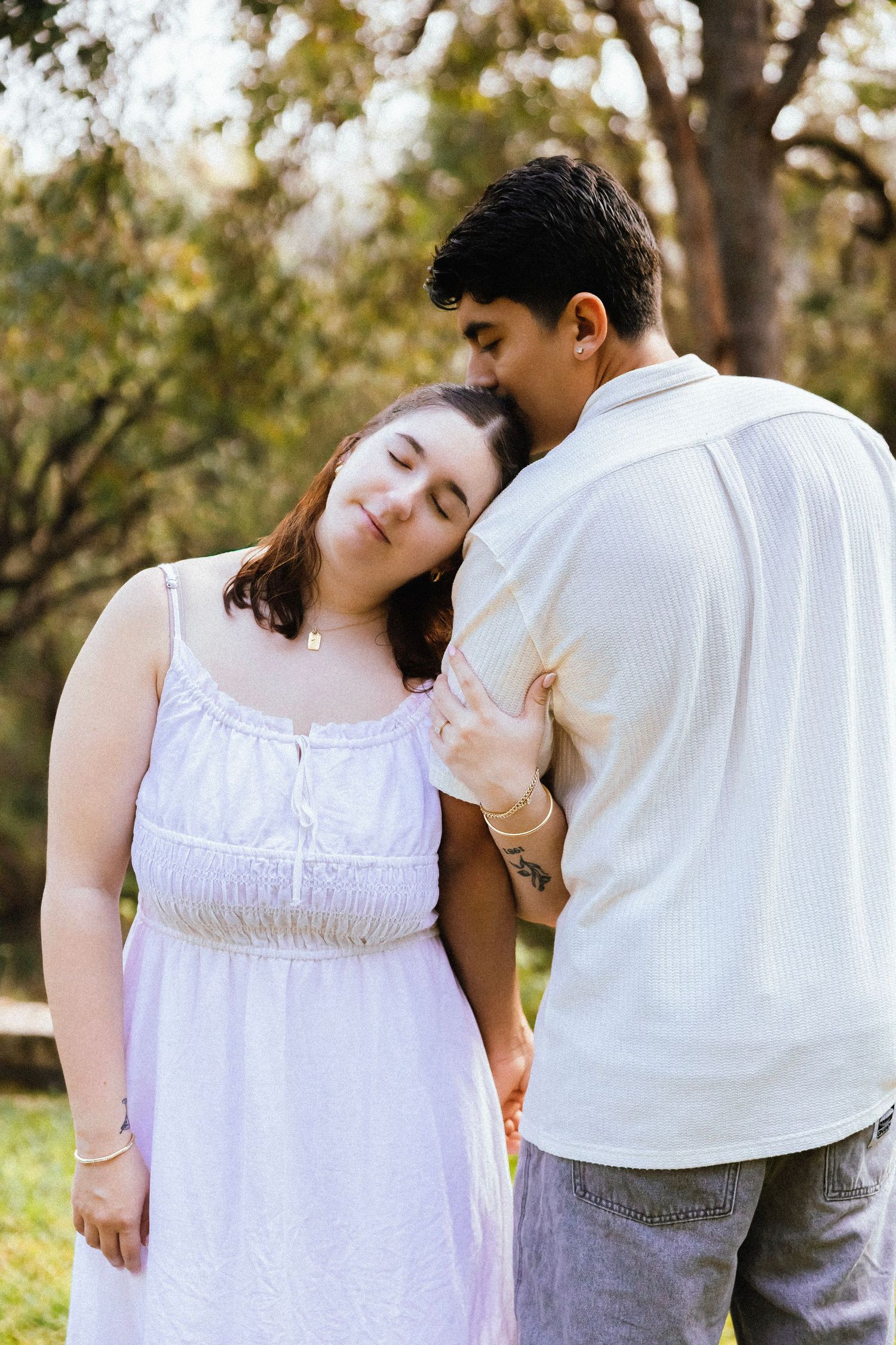 Woman in white dress leans head on partners shoulder smiling peacefully in a sunny park.