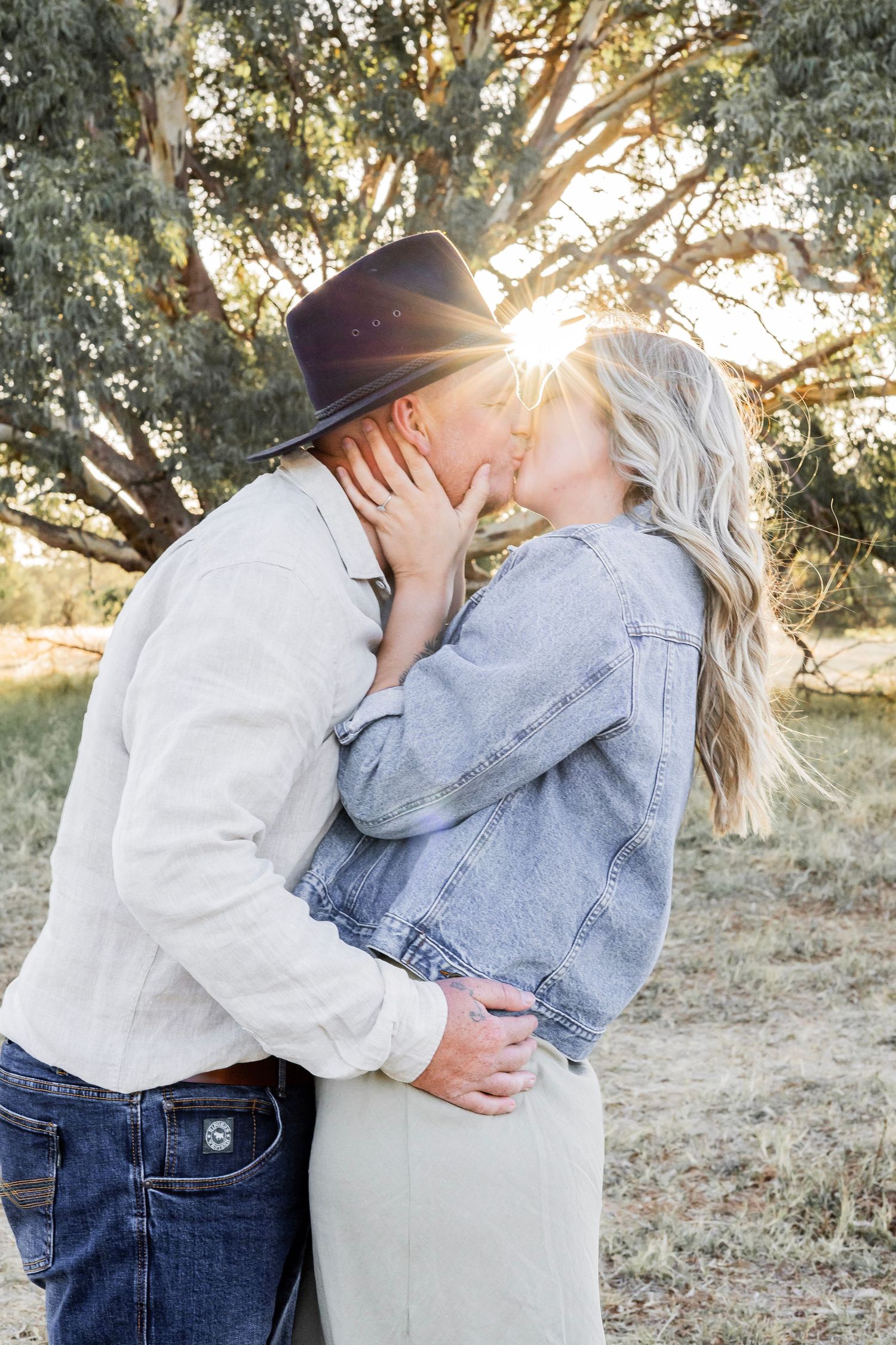A couple shares a kiss backlit by golden sunset light near trees in an outdoor field setting.