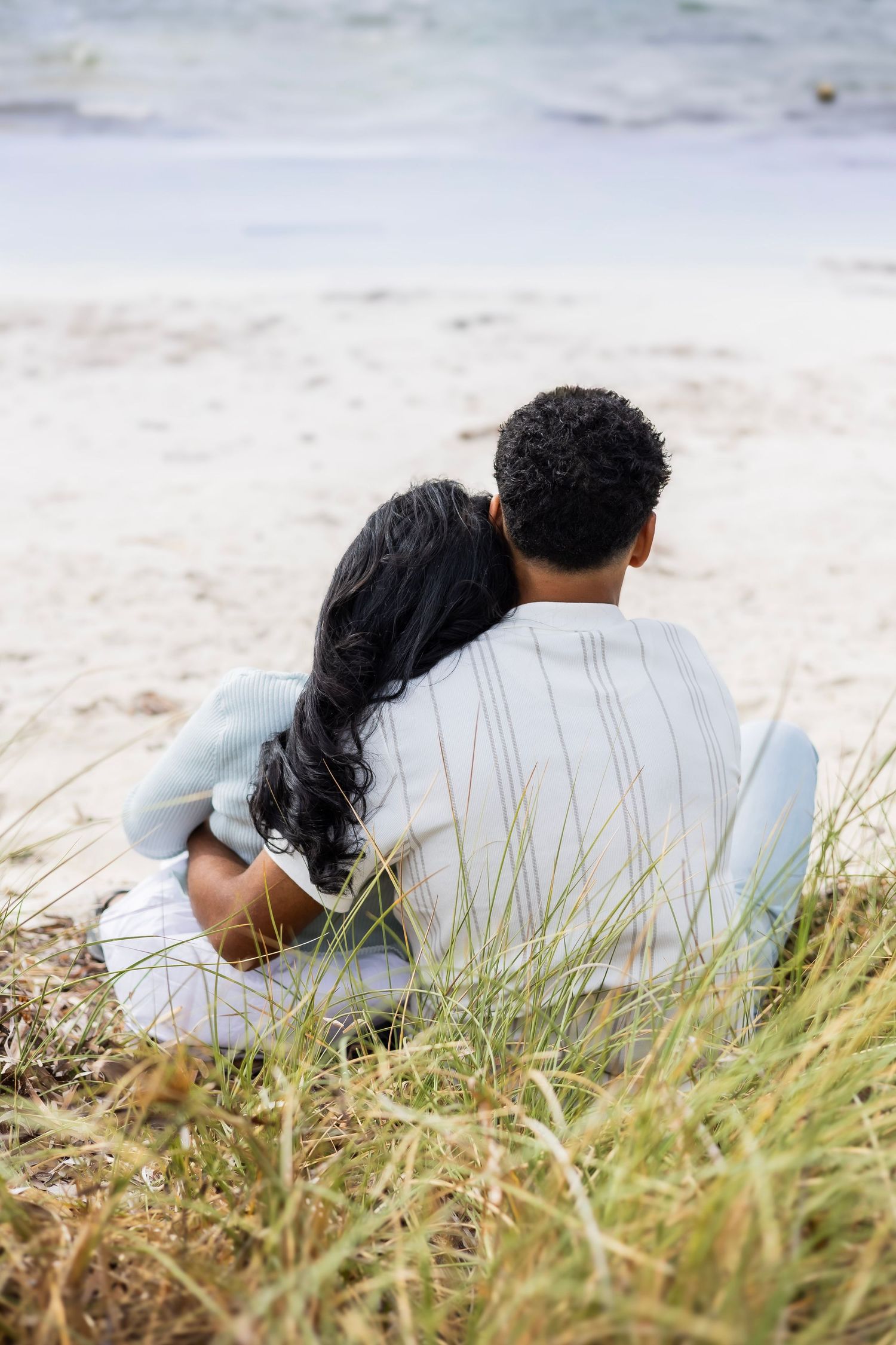 Couple sitting back-to-camera on beach sand, leaning on each other while overlooking the ocean.
