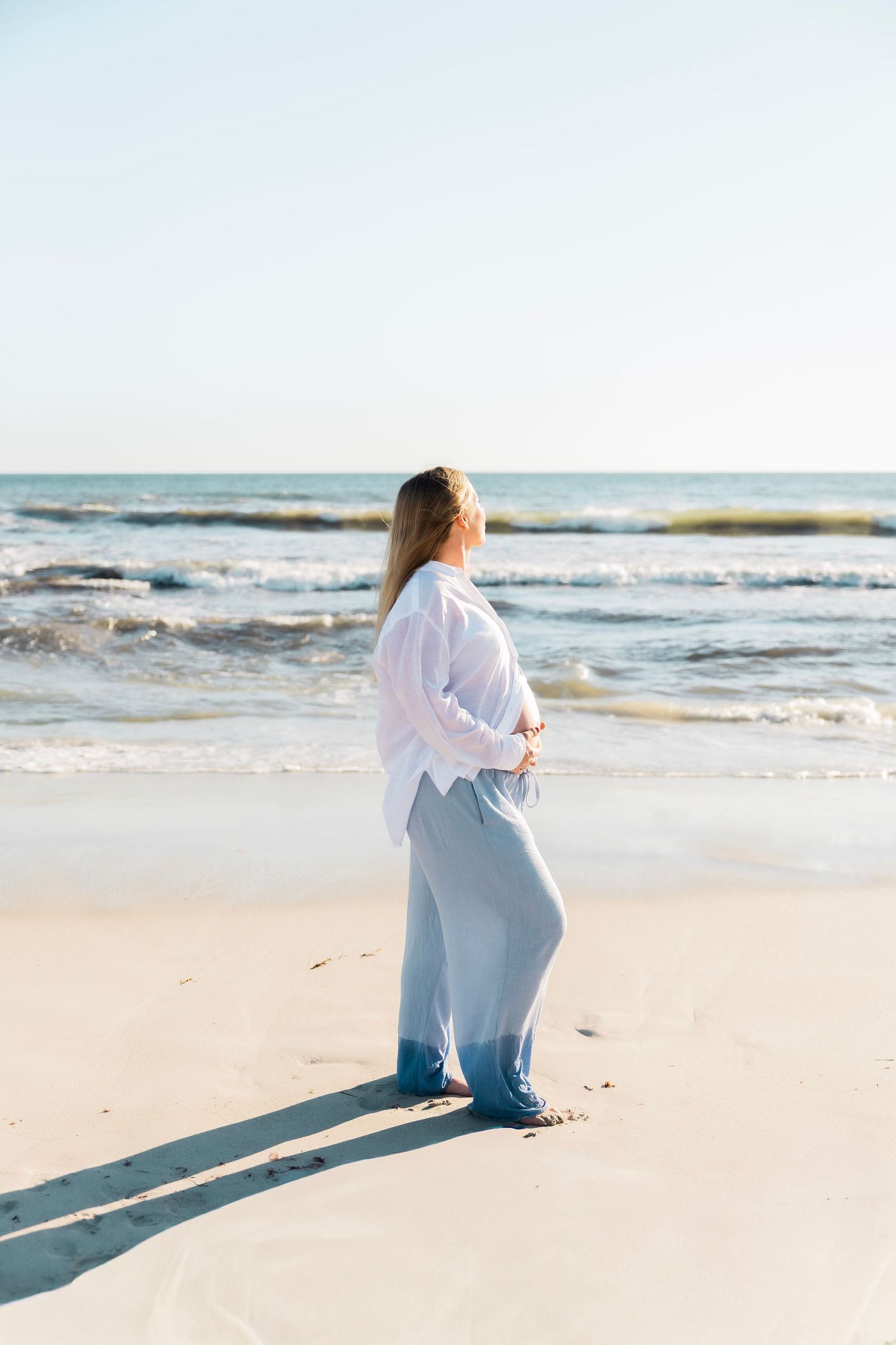 Pregnant woman in white shirt holding belly, standing on sandy beach facing the ocean.