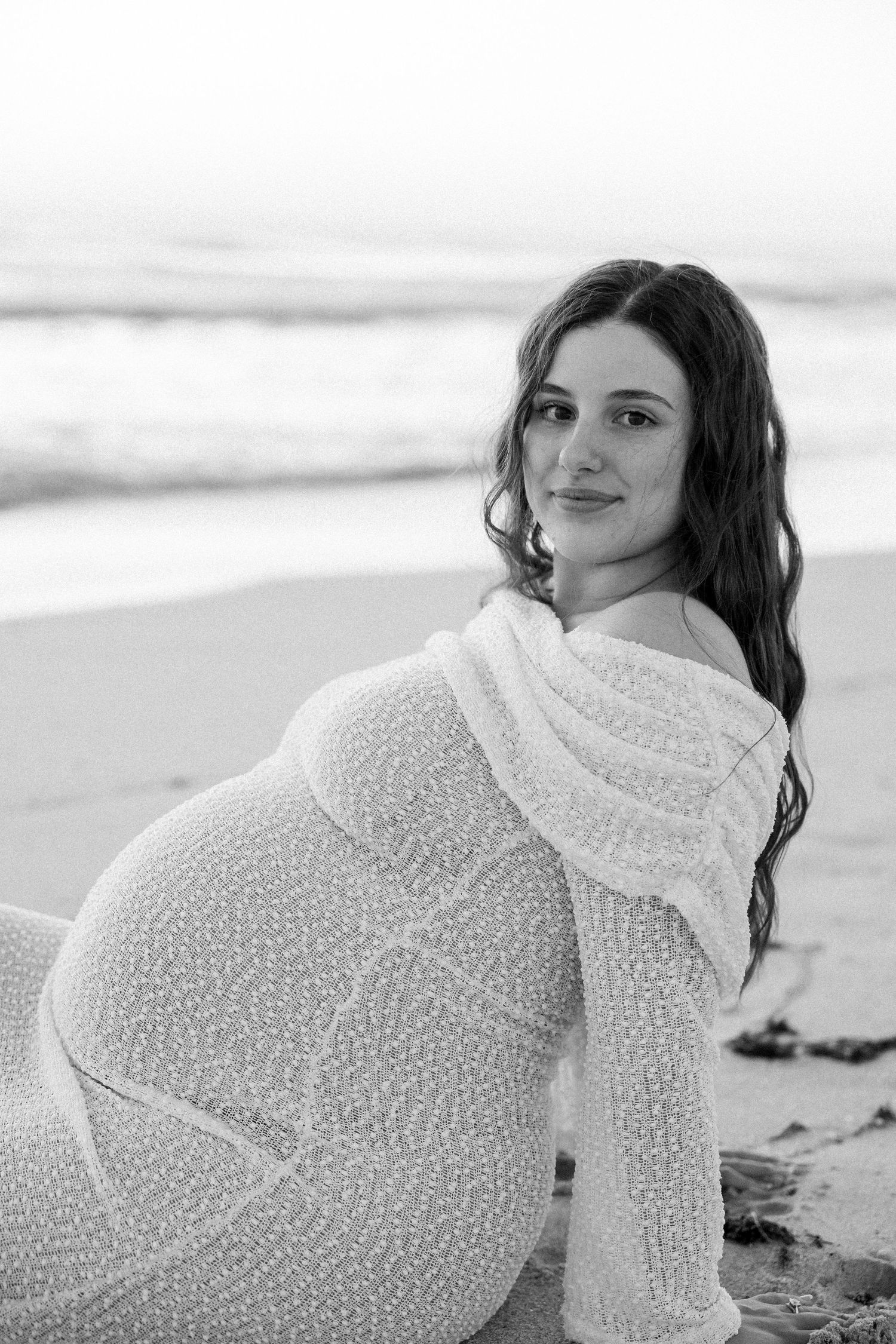 Black and white photo of smiling pregnant woman in white knit dress sitting on sandy beach by ocean.