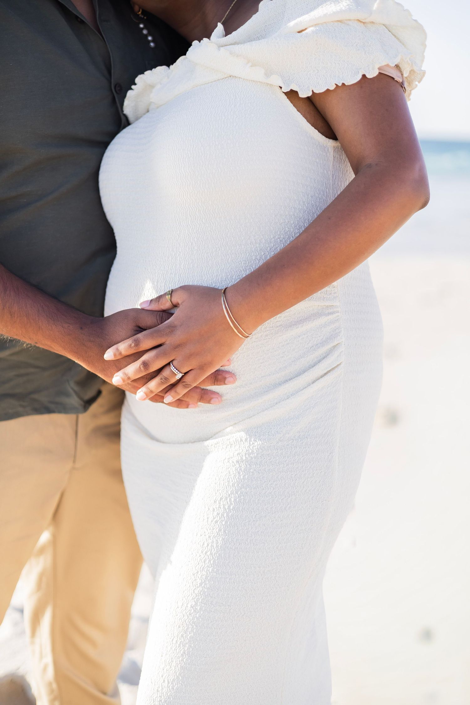 Pregnant woman in white dress with partner's hands on baby bump at sunny beach maternity photoshoot.