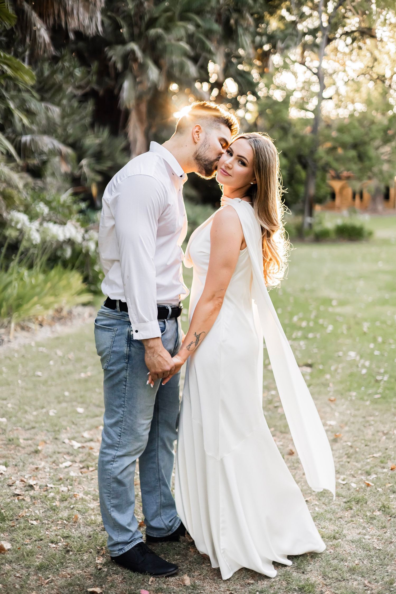 Bride in white gown and groom in jeans holding hands at golden hour in a tropical garden.
