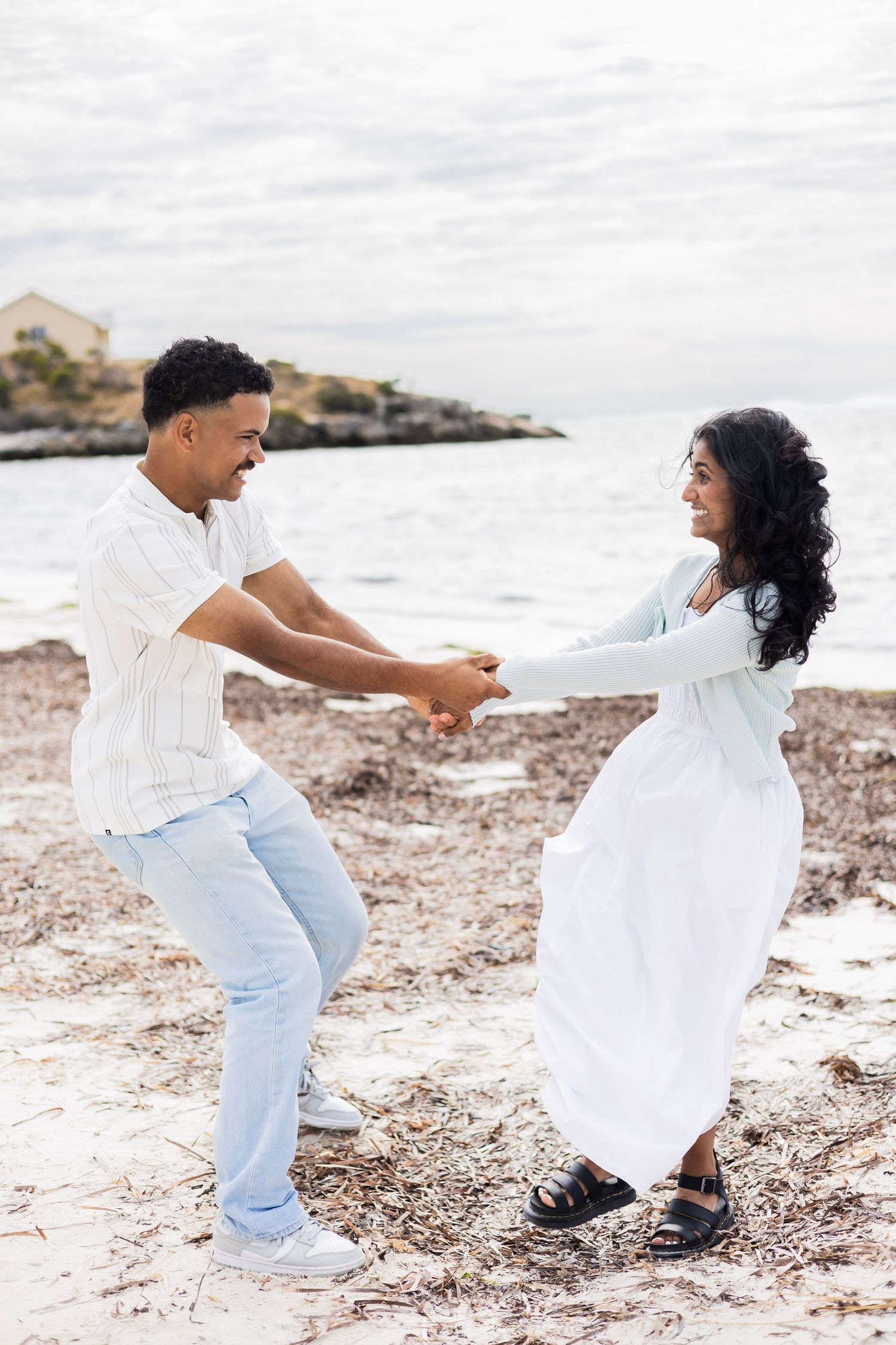 Couple dancing and spinning joyfully together on rocky beach shoreline with ocean backdrop.