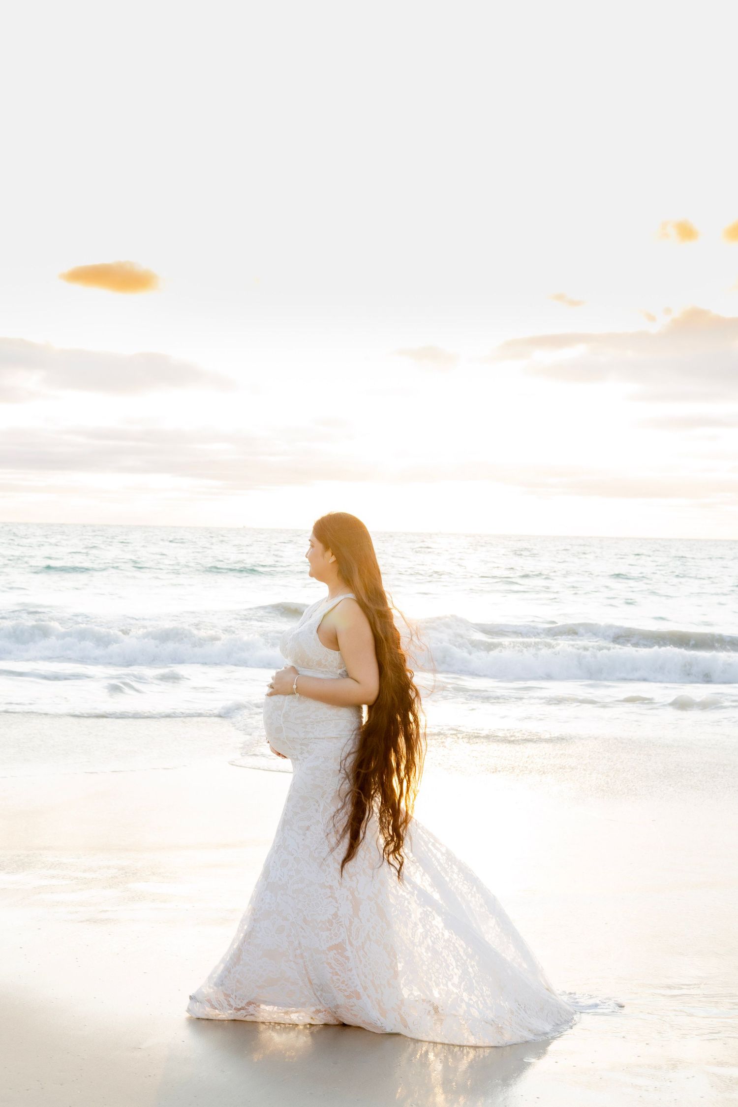 Glowing pregnant woman in white lace dress standing in ocean surf at golden sunset.