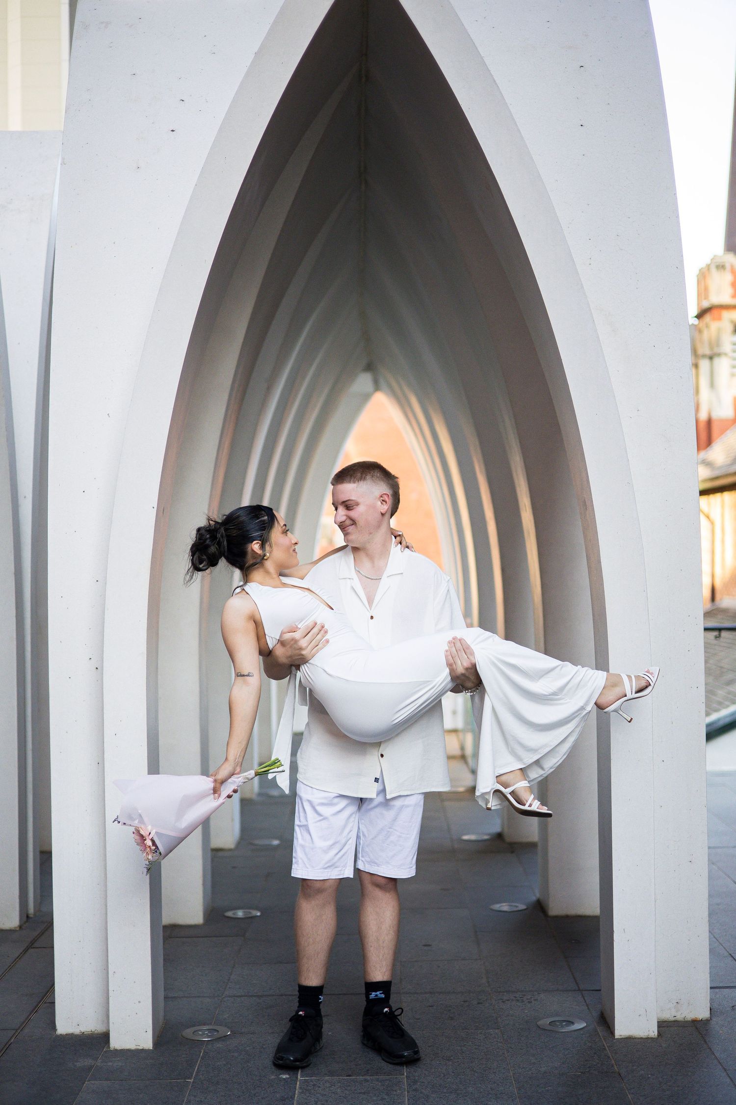 Couple sharing a joyful moment as man carries woman in white dress through white arched walkway.