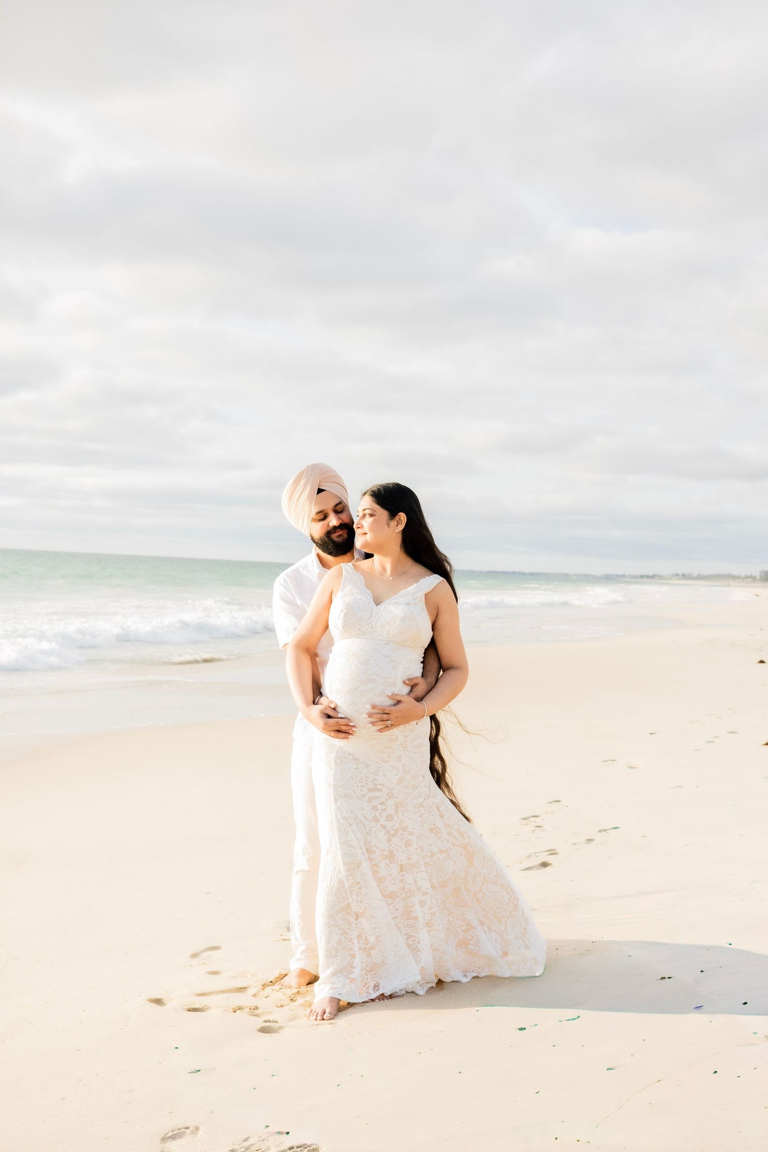 Pregnant woman in white lace gown embraced by partner in turban on a serene sandy beach.