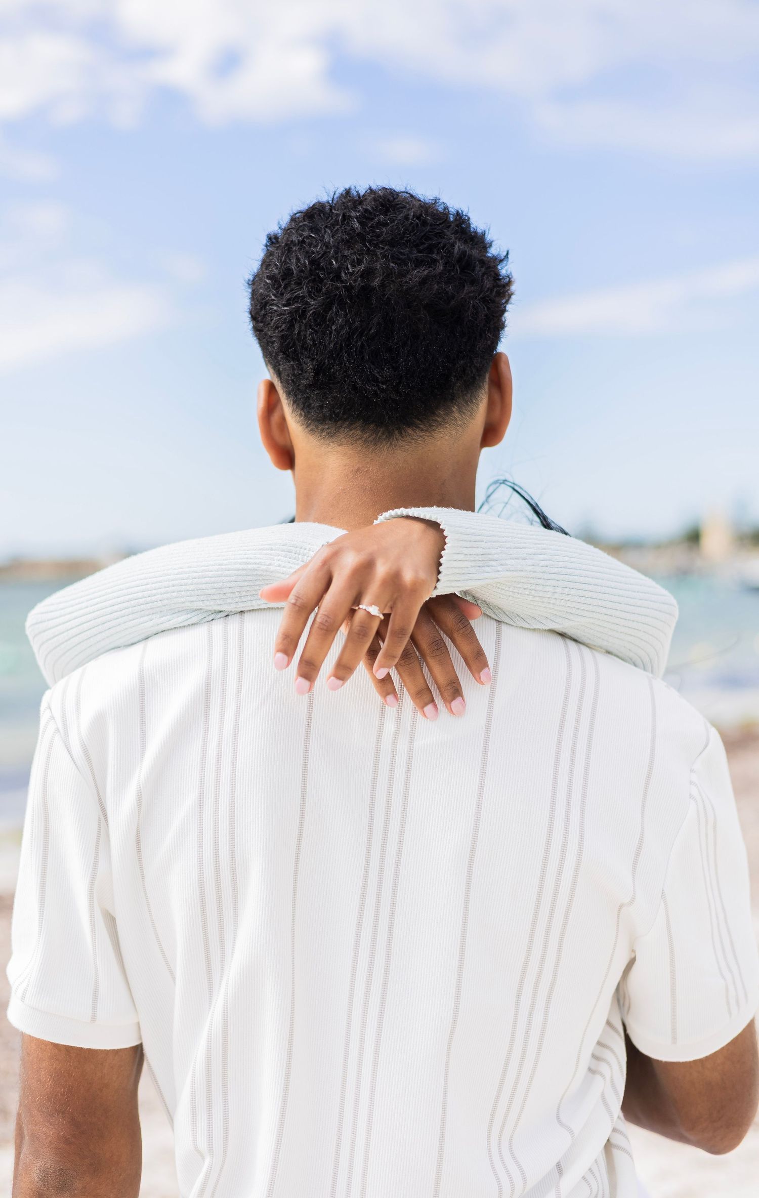 Close-up of a woman resting her hand with an engagement ring on a mans shoulder at the beach.