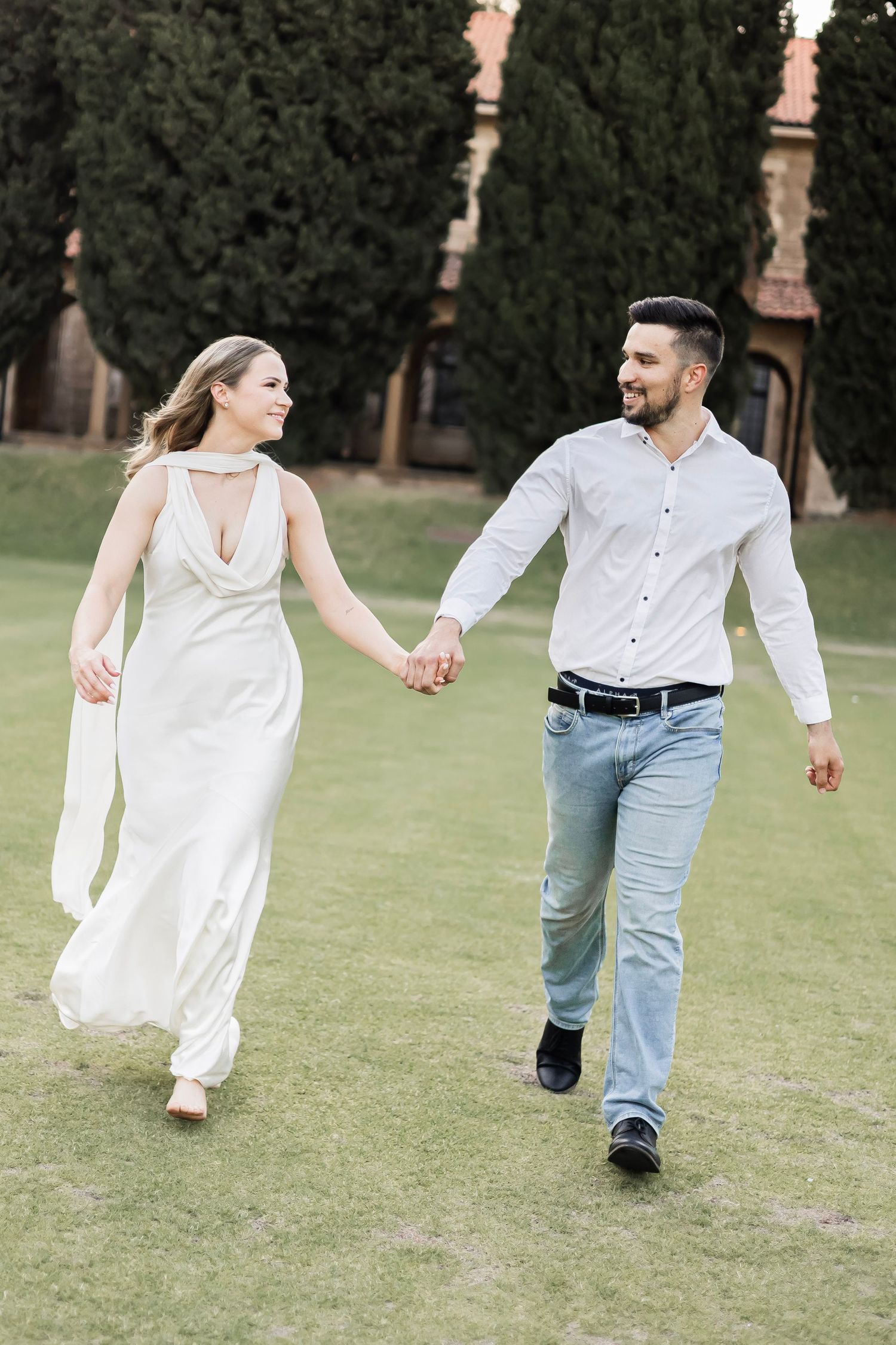 Happy couple holding hands and walking together on a green lawn near a historic building.