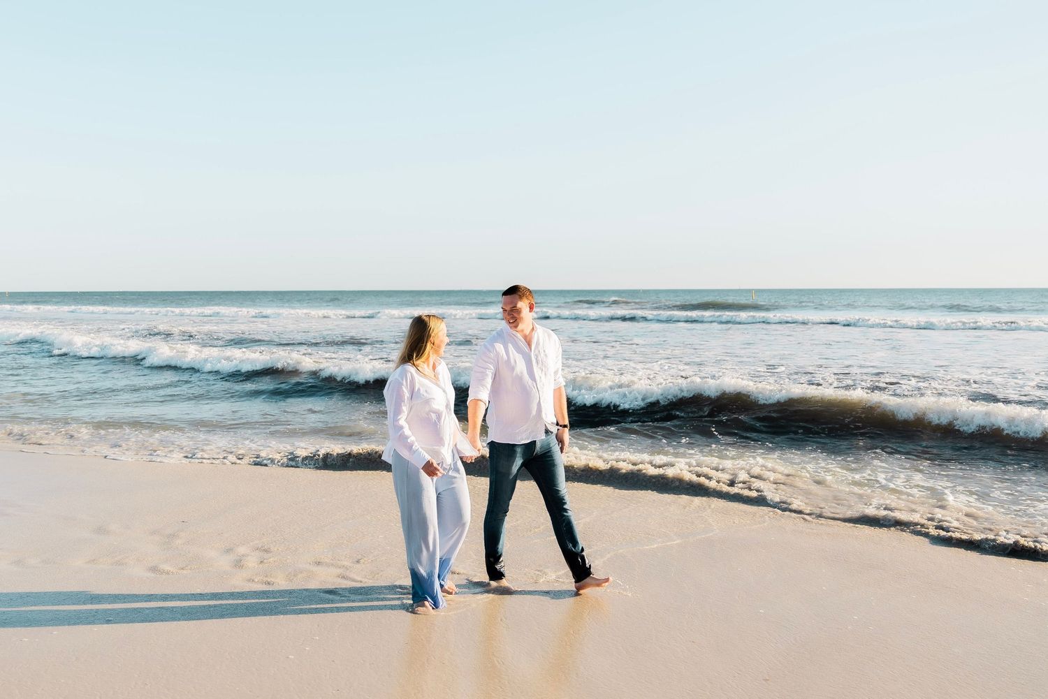 Couple holding hands walking on a sunny beach with ocean waves in the background.