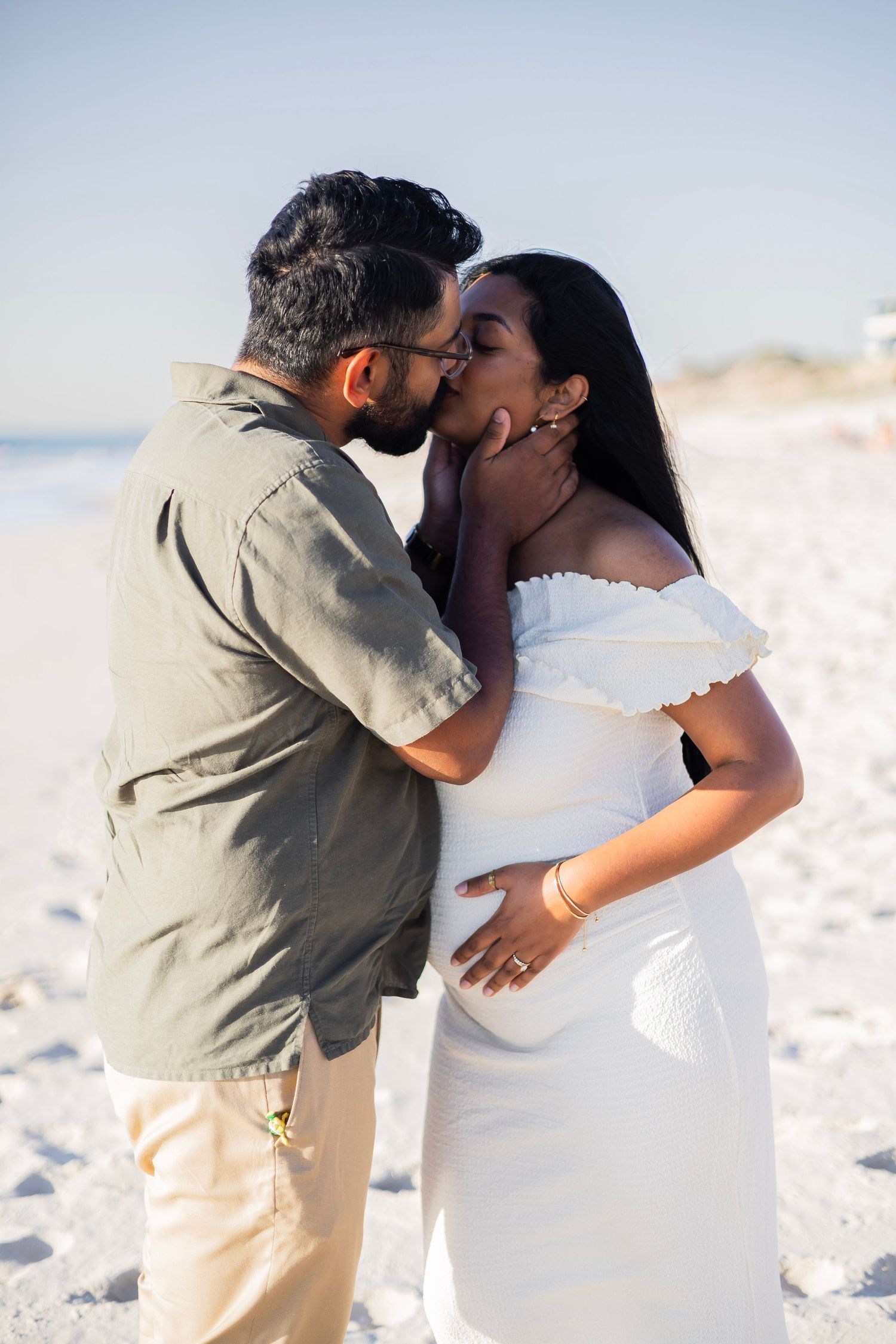 A couple shares a tender kiss on a sunny beach, with the woman in a white off-shoulder dress cradling her pregnant belly.