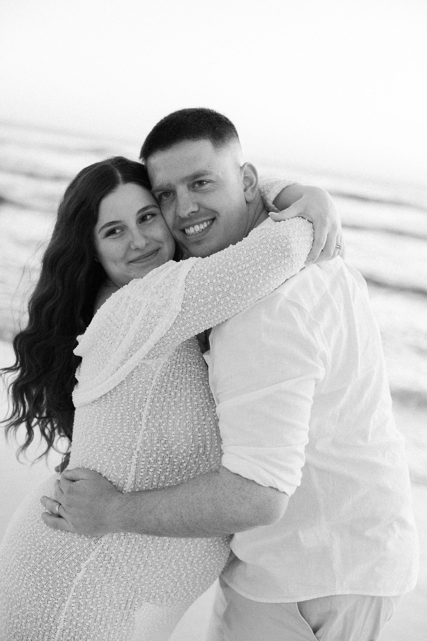 Happy couple hugging and smiling on beach in black and white maternity photo session.